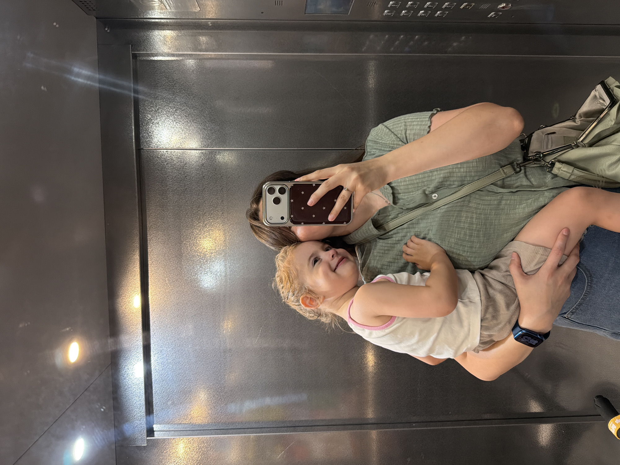 In this vertical, medium shot, a woman holds a young child in her arms while taking a selfie in what appears to be an elevator. The woman, seen from the chest up and reflected in the elevator's metal walls, wears a green, button-up shirt and holds a smartphone with a starry case. Her expression is partially obscured by the phone, but she seems to be smiling. The child, a toddler with blonde hair, is nestled in the woman's arms, facing the camera and smiling widely with their eyes closed. The child wears a white tank top with pink trim. The elevator's interior, with its brushed metal walls and bright, reflective surfaces, suggests an indoor, urban setting. Lights on the elevator ceiling cast a warm glow, creating reflections on the walls. The overall mood is warm and affectionate, capturing a candid moment between the two subjects. There is no visible text in the image.
