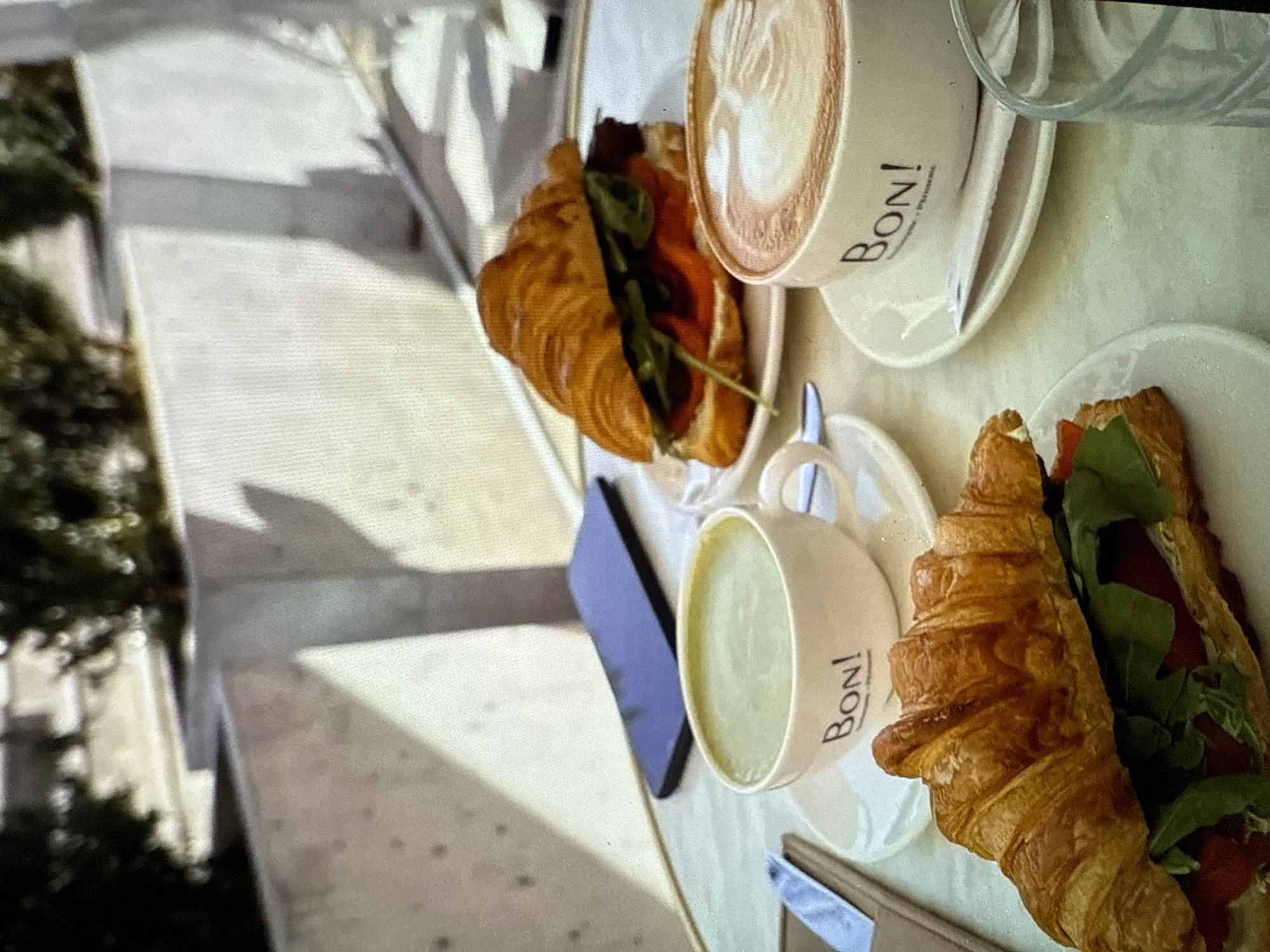 An overhead, medium shot captures a breakfast or brunch setting at an outdoor cafe. On a table with a light-colored, textured surface, two croissants filled with greens and red slices are arranged, one in the foreground and another further back. Two cups of coffee, one with latte art and the other containing a green beverage, sit on saucers with the "BON!" logo. A smartphone lies on the table near the cup with the green beverage. The background is blurred, suggesting an outdoor environment with plants and seating areas. The overall lighting suggests a bright, daytime scene.