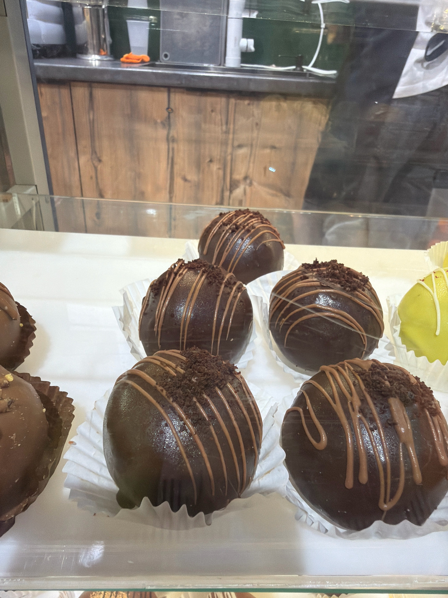 This image shows a close-up, eye-level view of several chocolate-covered spherical pastries displayed in a glass case. The pastries are dark brown, with lighter brown drizzles of chocolate on top and sprinkled with dark crumbs. They are presented in white paper liners. In the background, there is a wooden paneling surface. To the far right, a yellow pastry is partially visible. The scene appears to be inside a food establishment, likely a bakery or confectionery. The image does not contain any people or any specific identifiable landmarks that would indicate the city of Pavlodar, Kazakhstan.