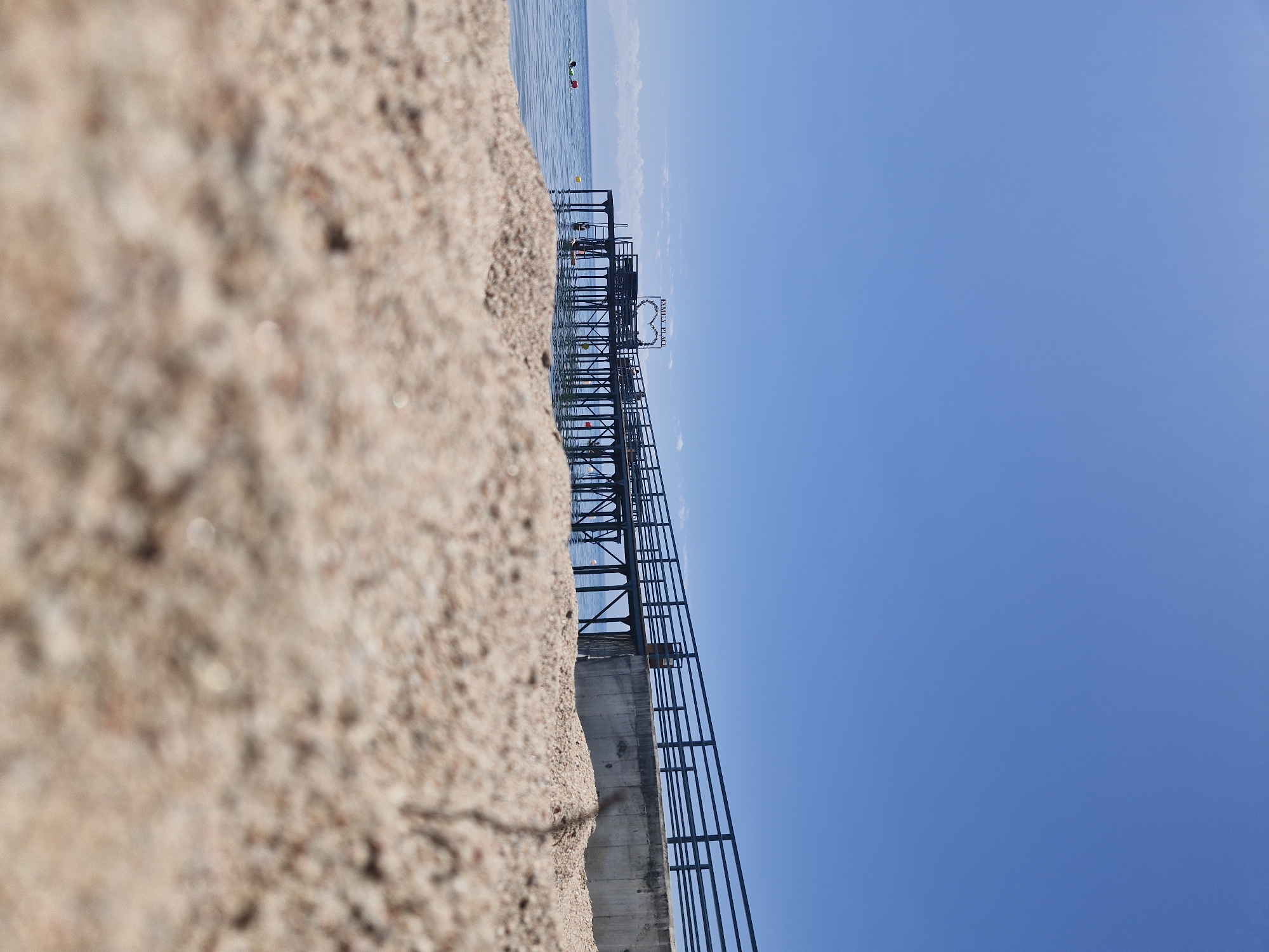 A pier extends over a body of water under a clear blue sky. The foreground is occupied by sand, out of focus. The pier has a railing and is supported by numerous vertical posts. A sign shaped like a heart with the words "FAMILY PLACE" is visible on the pier. There are several people in the water near the shore and on the pier. In the distance, mountains are faintly visible on the horizon. The scene appears to be a beach resort or a recreational area.