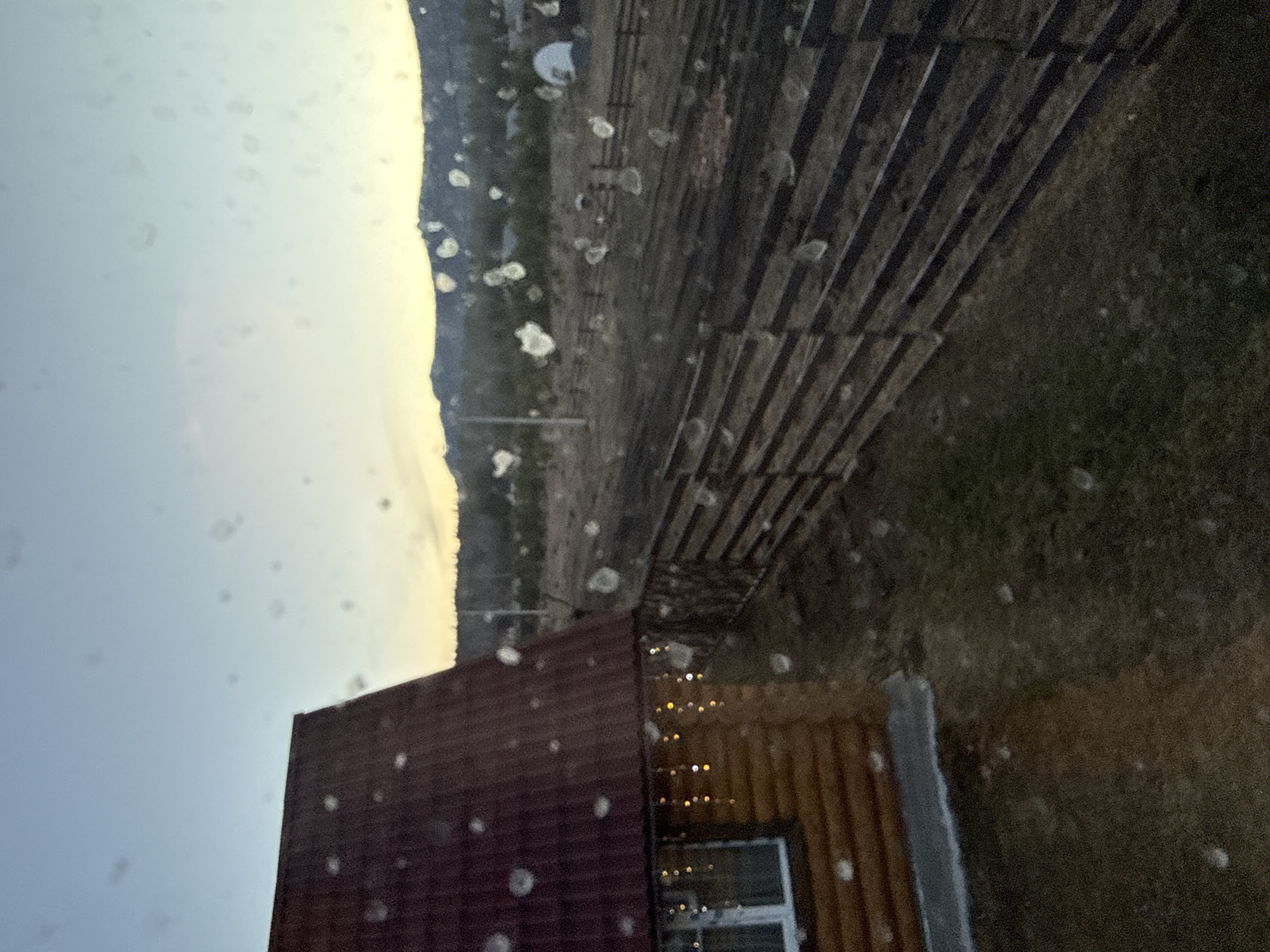 The image depicts an exterior scene in Tyulyuk, Russia. It shows a building with a red-tiled roof and wooden siding. A small window with a white frame is visible. Adjacent to the building, there's a wooden structure, likely a fence or retaining wall, with a slatted design. The background includes a landscape with trees and possibly a distant mountain range. The sky appears overcast. There are spots or droplets visible, potentially on a window or lens, indicating it might be raining or wet conditions. The scene lacks human presence or any explicit action.