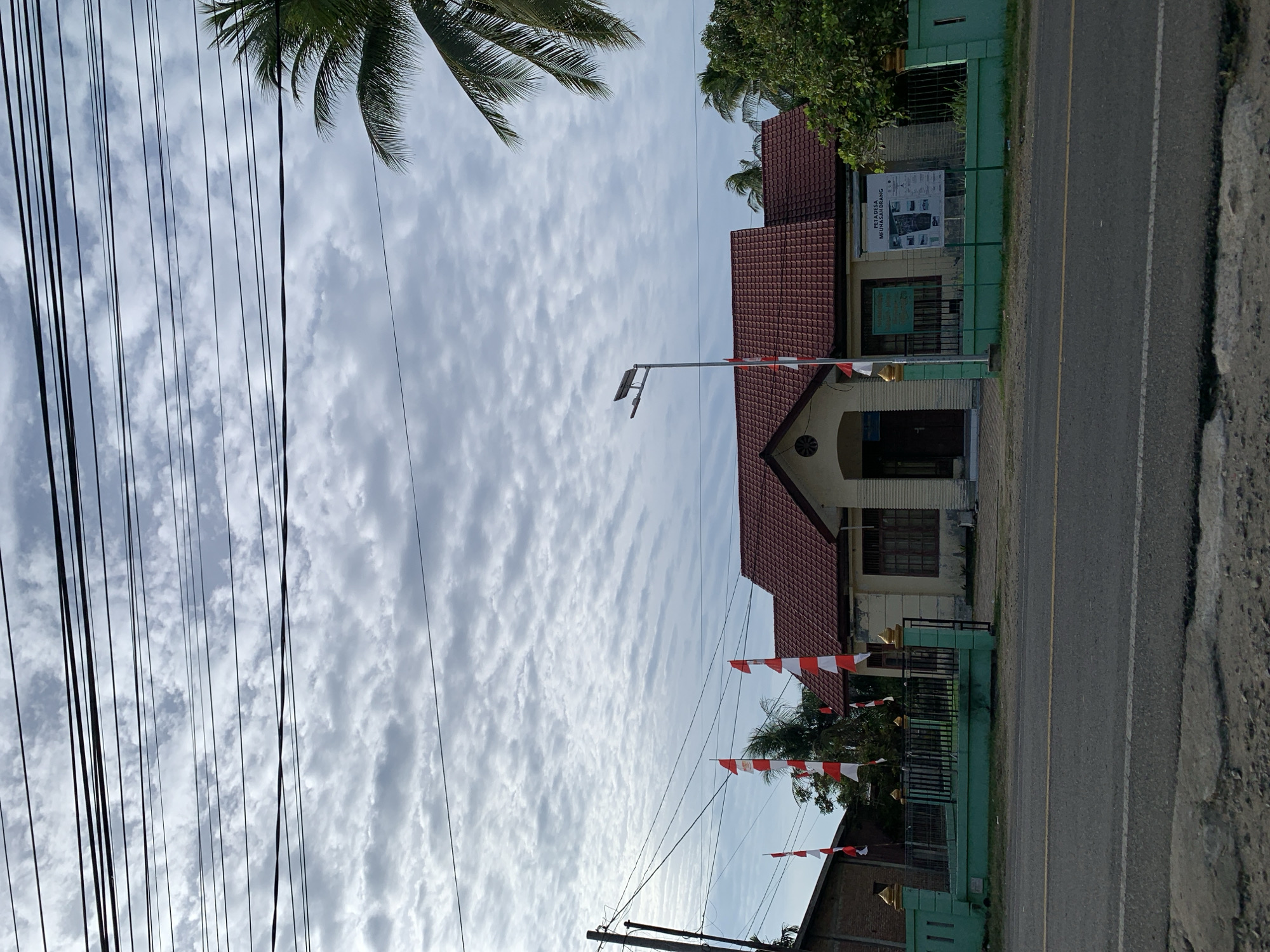 The image captures a building in Meunasahlhok, Indonesia, with Indonesian flags displayed. The building appears to be a public facility, possibly a village office, given the "PETA DESA MEUNASAHLHOK" (Village Map Meunasahlhok) poster visible on its exterior wall. The building has a red tiled roof and light-colored walls, with a green fence surrounding the property. The scene is set along a road, with power lines and a street lamp visible. A palm tree is also present on the right side of the image. The sky is cloudy, suggesting overcast weather. There are no people visible in the image.