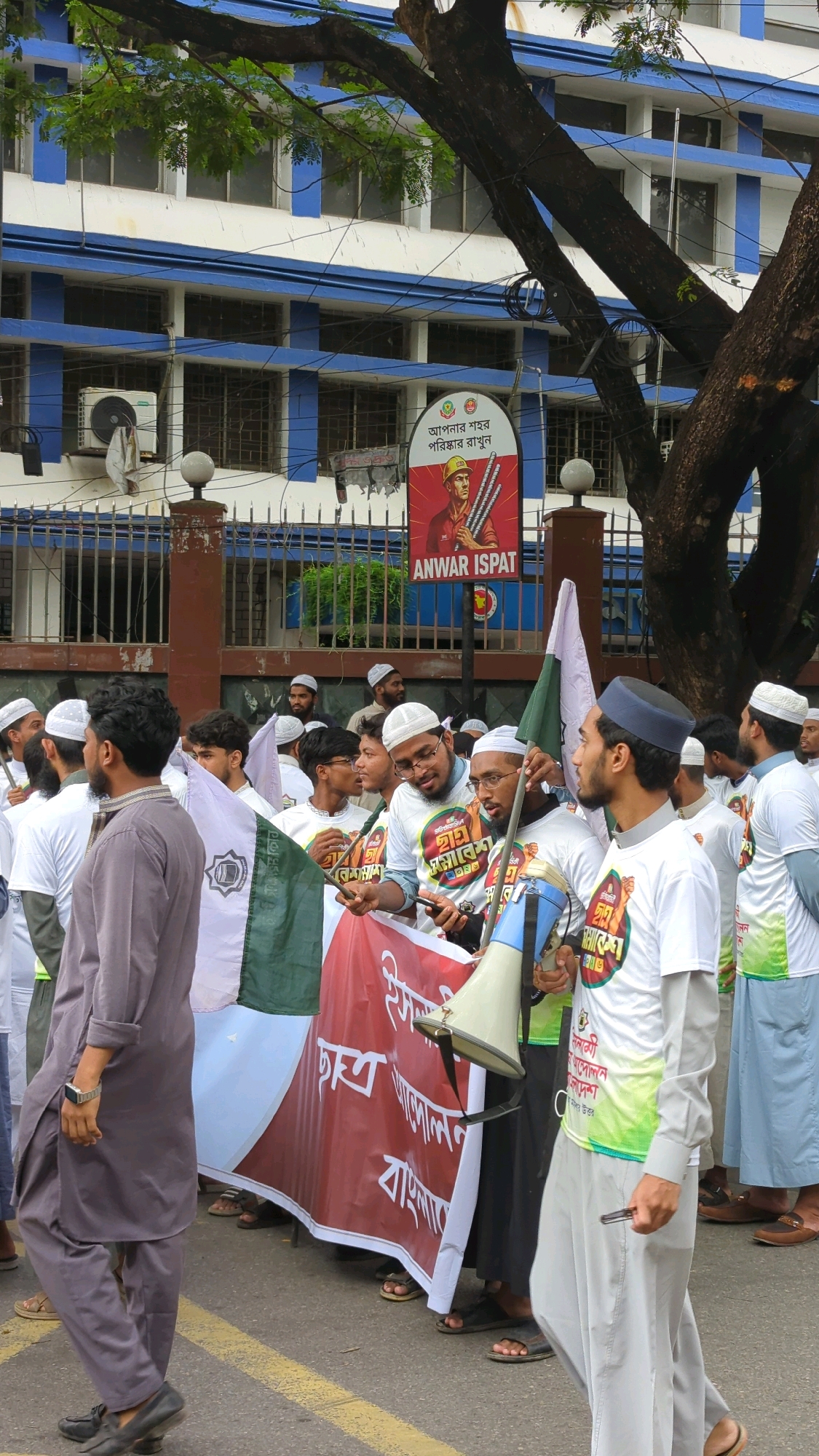 In Dhaka, Bangladesh, a group of men wearing Islamic skullcaps and some with white t-shirts with colorful text are seen walking on a street. They are carrying flags and a large banner with Bengali text. In the background, a white and blue building with barred windows stands. A large tree partially obscures the view of the building. A sign with an image of a worker holding steel rods and the text "ANWAR ISPAT" is also visible. The men appear to be participating in a procession or protest. The ground is paved and a yellow line indicates a lane divider.