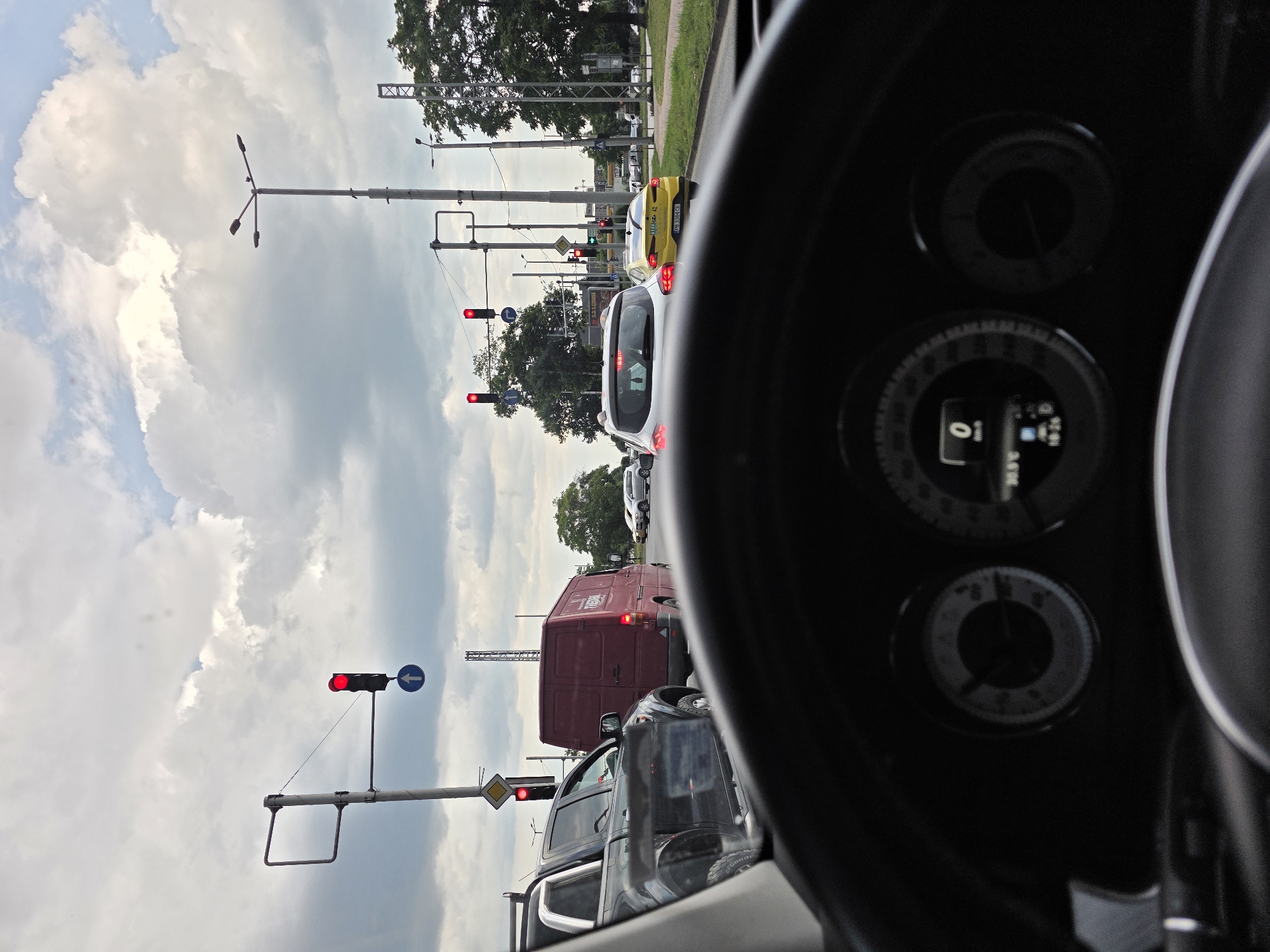 The view from inside a car, focusing on the steering wheel and dashboard, reveals a traffic situation. The speedometer reads zero, indicating the vehicle is stationary. Through the windshield, several vehicles are stopped at a red traffic light. A red van is prominent in the mid-ground, with a yellow car further ahead, partially visible on the right side of the frame. Traffic lights and support poles are visible along the roadside, against a backdrop of cloudy skies and some trees. The location is Plovdiv, Bulgaria, based on the appearance of road signs and vehicle license plates.