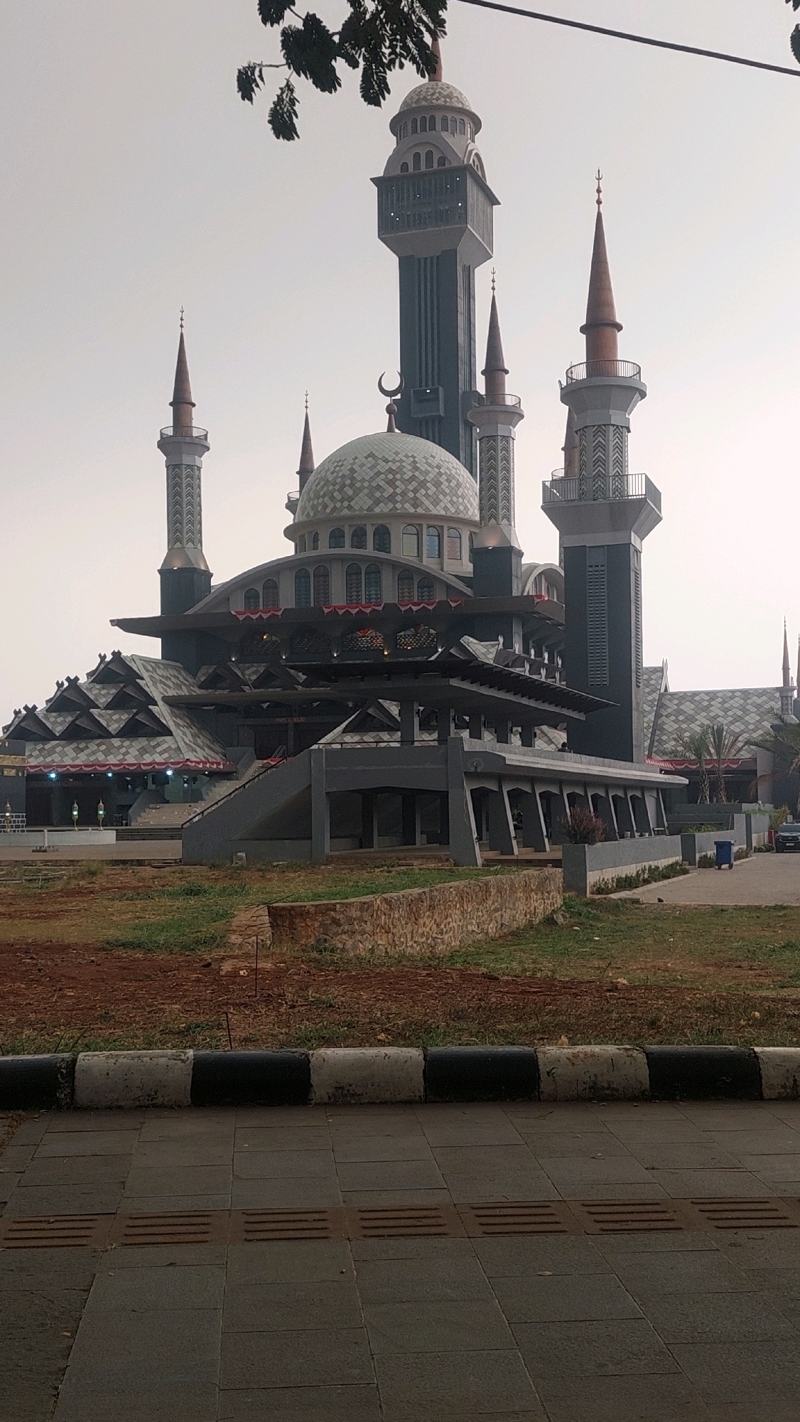 The image shows the exterior of the Cibinong Grand Mosque, also known as Masjid Agung Cibinong, in Cibinong, Indonesia. The mosque is characterized by its multi-level architecture, featuring a prominent central dome, several tall minarets of varying designs, and a complex roof structure with geometric patterns. The main building is dark gray and gray, with some white and light brown accents. Red and white decorative banners are visible along some of the building's edges.
In the foreground, a paved walkway with textured tactile paving for the visually impaired runs parallel to a low stone wall and a grassy area. Beyond the mosque, a few trees are visible, and the sky is overcast. The overall lighting suggests it is daytime, likely a hazy or cloudy day. There are no people clearly visible in the image, nor any discernible actions or interactions. The scene depicts a static view of the mosque and its immediate surroundings.