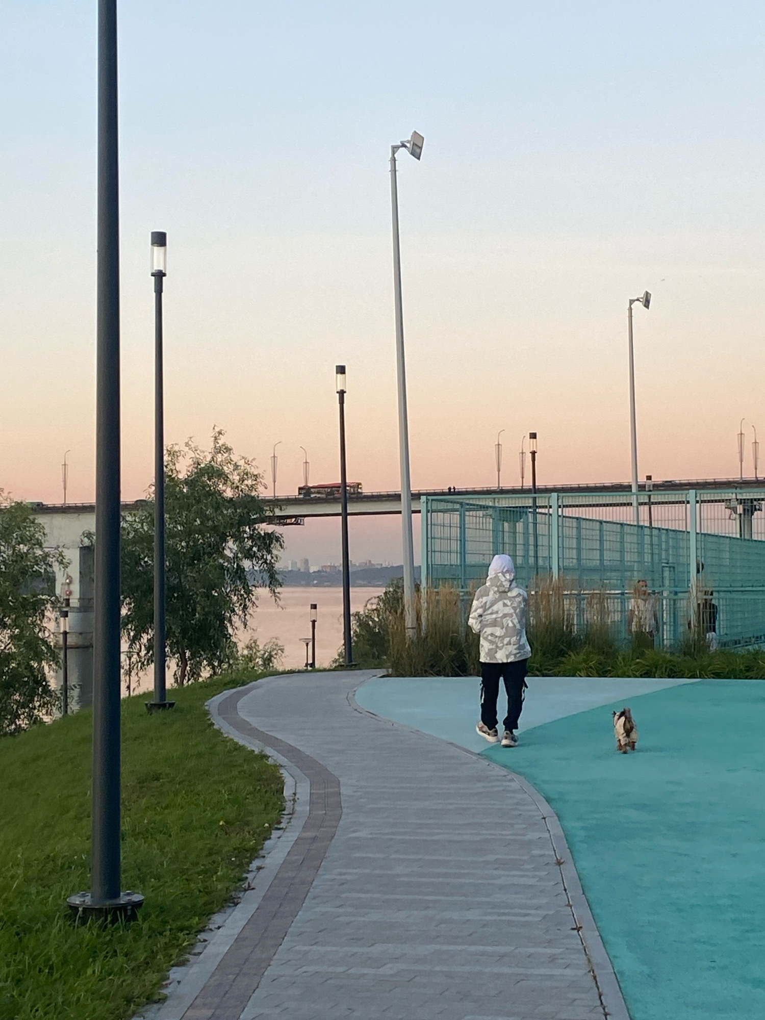 In Perm, Russia, a person in a camouflage jacket and white head covering walks along a paved pathway next to a grassy embankment. A small dog with light brown fur follows behind. The pathway curves away from the viewer, leading towards a turquoise-colored recreational area. In the background, a wide river is visible, crossed by a bridge. A bus is traveling on the bridge. Several lampposts are present along the pathway and near the river. A light blue sky with a soft pink hue suggests it is either dawn or dusk. The scene appears to be a public park or waterfront area.