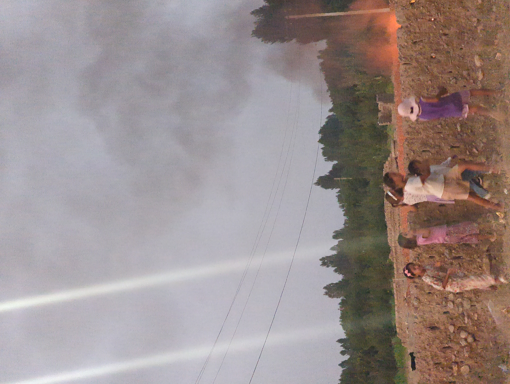 In the village of Uch-Dobo, Kyrgyzstan, a group of five children, appearing to be between the ages of five and ten, are gathered in an outdoor setting. They are standing on a dirt path next to a low, rough-hewn stone wall. Behind the wall, dense green trees form a treeline. In the distance, smoke billows upwards into a hazy, overcast sky, with visible beams of light piercing through the clouds. Flames are visible in the lower right corner, suggesting a fire in the background. The children, dressed in casual clothing, are interacting with each other in what appears to be a spontaneous moment. Some are looking towards the fire and smoke, while others are engaged with one another. The overall atmosphere of the image is one of curiosity and a slight sense of unease due to the visible fire and smoke.