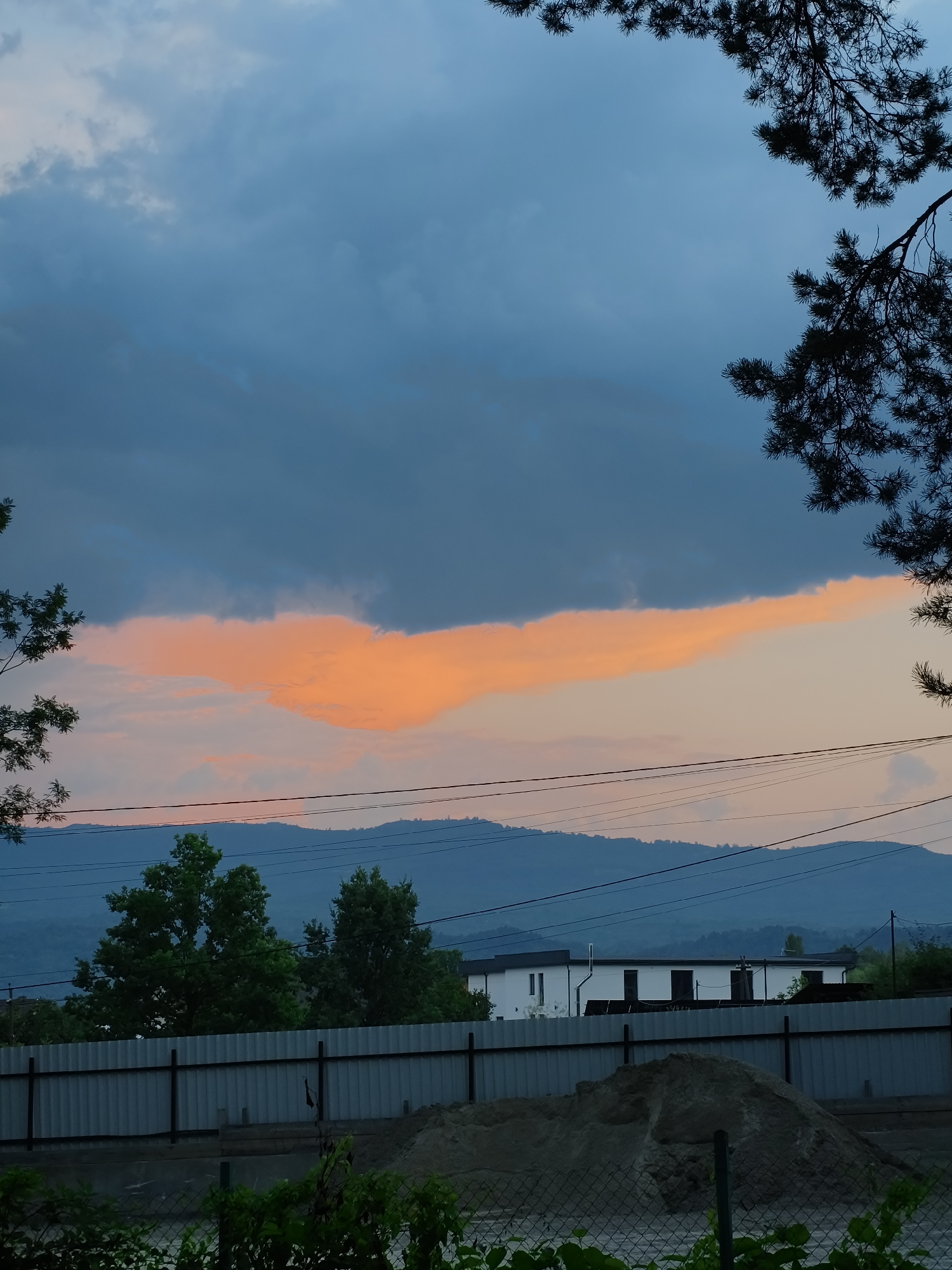 The image depicts a scenic view of the sky and landscape in Solotvyno, Ukraine. The sky is filled with dramatic clouds, with a striking band of orange-pink clouds cutting horizontally across a darker blue expanse. Silhouetted against the sky are the dark, feathery branches of pine trees in the foreground, framing the scene. In the middle ground, a range of blue-hued mountains forms the distant horizon, with power lines crisscrossing the sky above them. Closer still, a row of green trees stands tall, partially obscuring a modern white building with dark windows. A grey corrugated metal fence runs across the lower portion of the image, with a pile of sand and construction materials visible behind it. The overall impression is one of a natural landscape with human elements, likely captured at dusk or dawn due to the vibrant cloud colors.