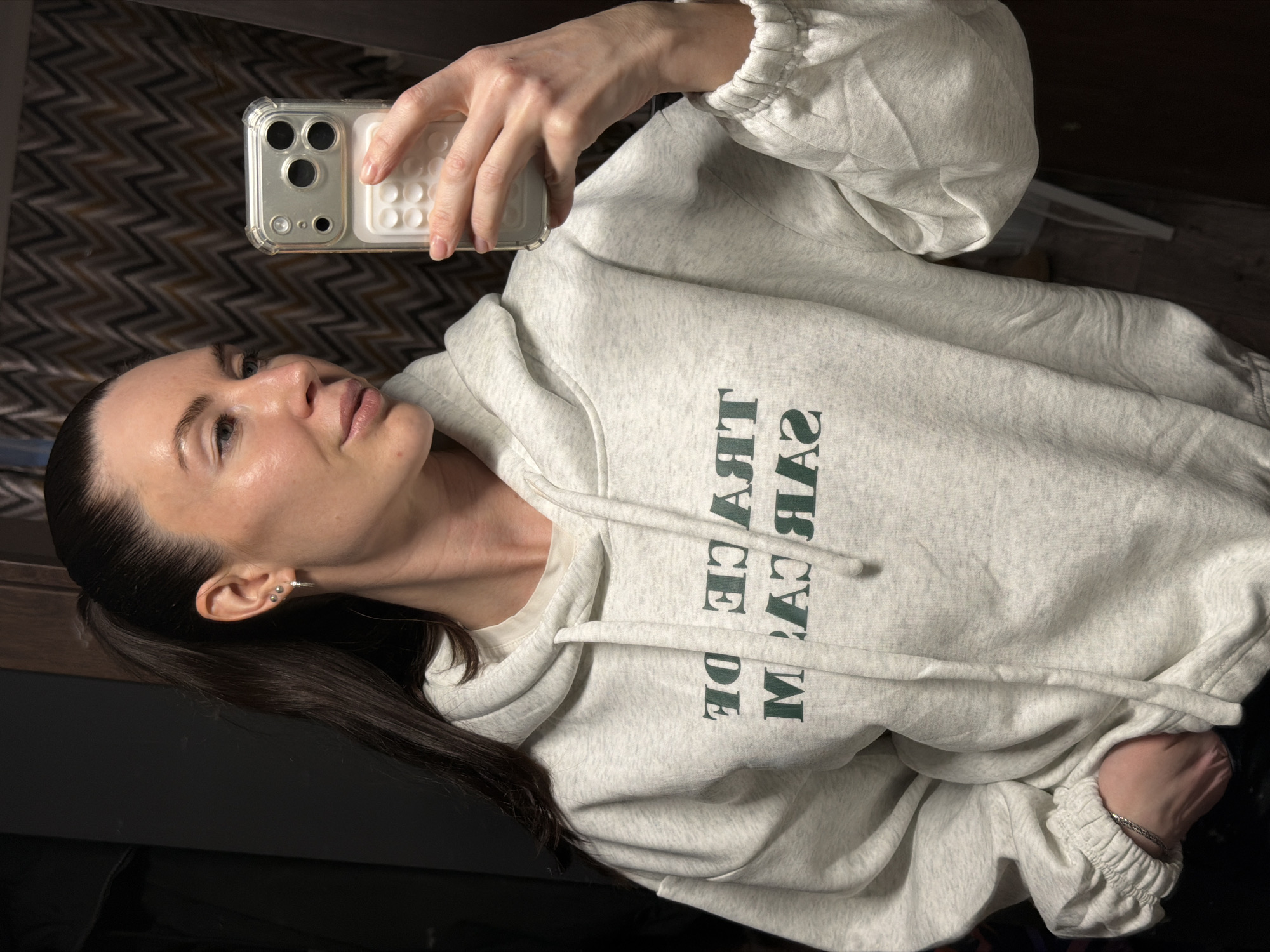 A woman is taking a selfie indoors. She is holding a smartphone in her right hand, and her face is visible in the frame. She is wearing a light gray hoodie with dark green lettering, and her hair is dark and pulled back into a ponytail. She is wearing earrings in her left ear. The background is dark and indistinct. It is not possible to determine the location as Perm, Russia, based solely on the visual information provided.
