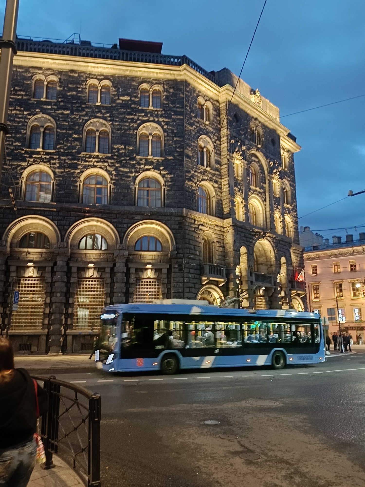 The image shows a blue bus driving on a wet street in front of a large, ornate stone building. The building has many arched windows and is illuminated by warm yellow lights. The sky is dark blue, suggesting it is dusk or early evening. Several people are visible inside the bus, and a few people are standing on the sidewalk to the right of the bus. A woman wearing a black hoodie and jeans is walking away from the camera on the left side of the image, near a black metal fence. The scene appears to be in a city, likely Saint Petersburg, Russia, given the architecture and the presence of a public transportation bus.