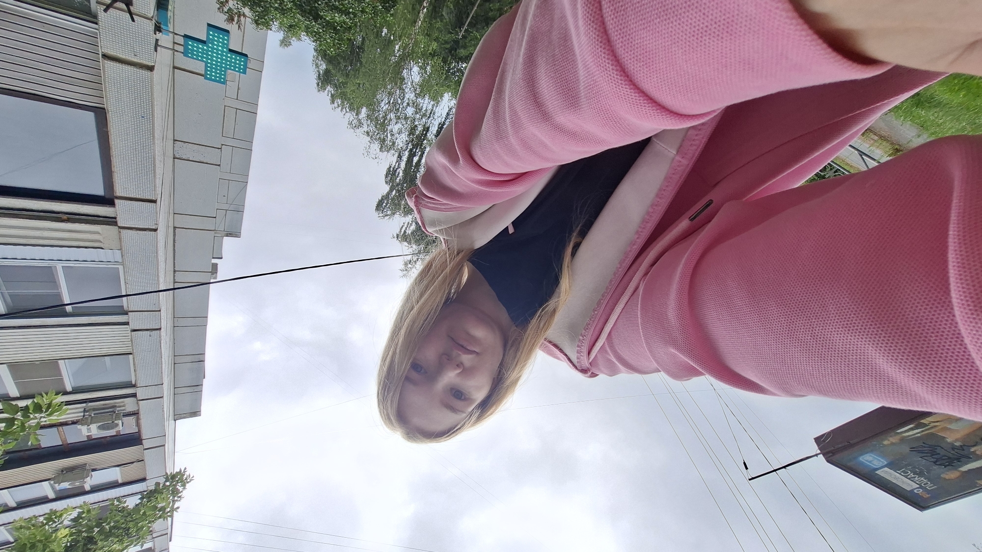 A young woman with blonde hair, wearing a pink hooded jacket over a black shirt, takes a selfie outdoors. She is looking directly at the camera with a neutral expression. The background shows a building with windows and air conditioning units, a green plus-sign-shaped sign, trees, power lines, and a billboard with text in Russian that appears to advertise a podcast. The photo is taken from a low angle, looking up at the woman and the surroundings. The sky is overcast. This scene is likely located in the city of Chaikovsky, Russia.