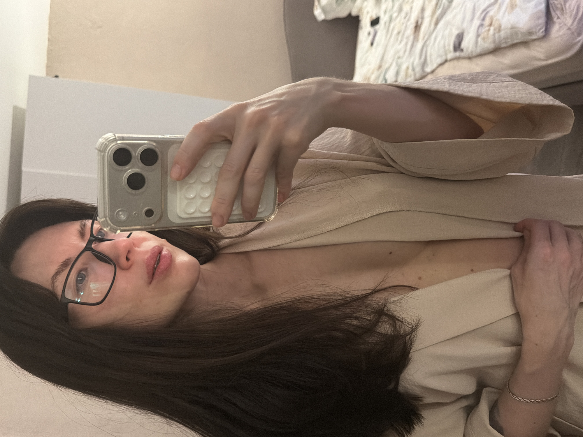 A woman with dark hair and glasses is taking a selfie with a smartphone. She is wearing a beige robe and has her hand up holding the phone. The background includes a white wall, a bed with patterned bedding, and potentially part of a headboard. The location information (Perm, Russia) cannot be confirmed with the visual evidence.