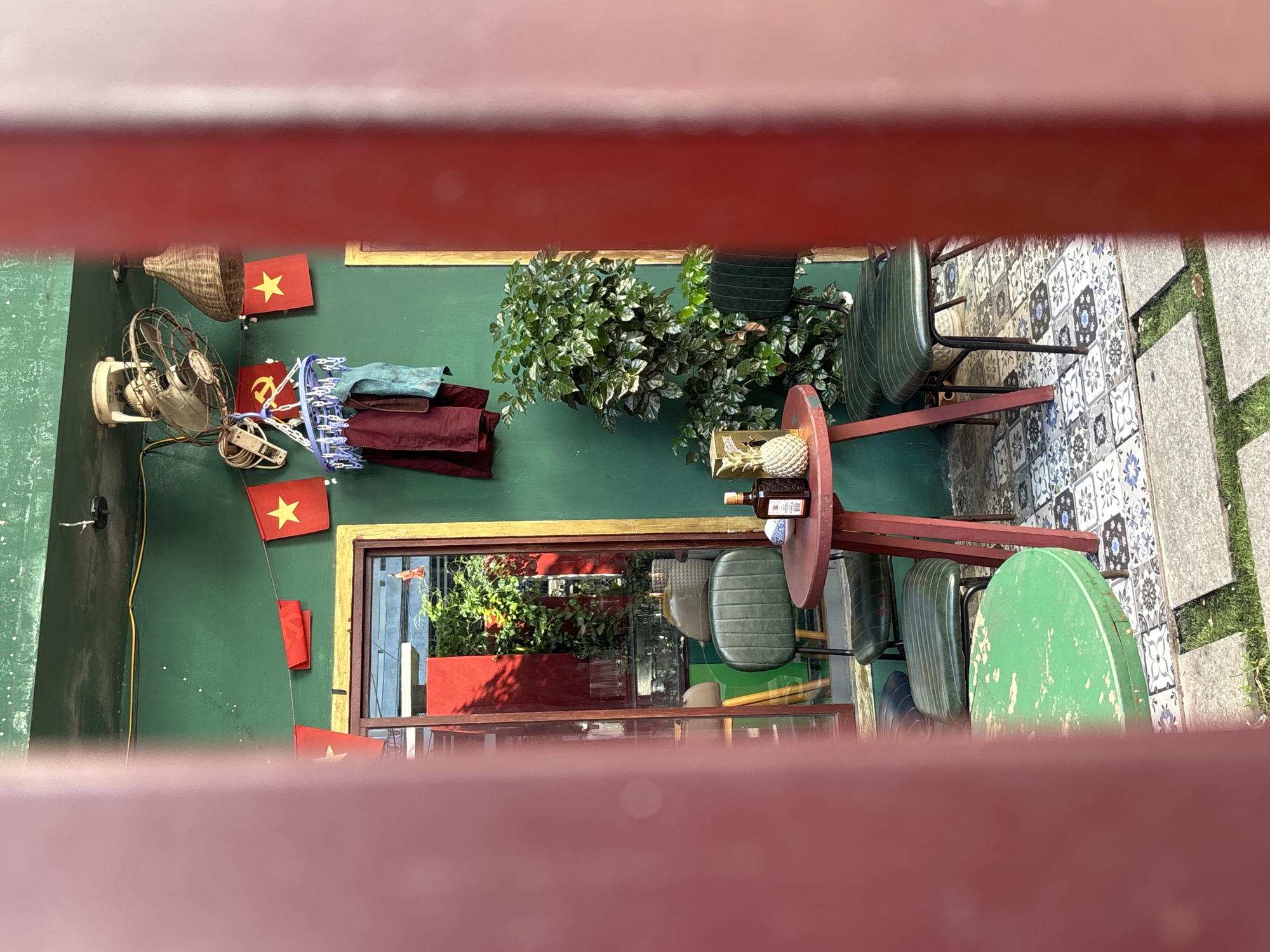 A street-level outdoor seating area, likely a cafe or restaurant, is captured through a red-barred foreground. The walls of the building are painted a vibrant green. A string of small red flags with yellow stars, reminiscent of Vietnamese national flags, is strung across the upper portion of the wall. A vintage electric fan is mounted on the ceiling, and a woven light fixture hangs nearby. Clothes are hung on a drying rack suspended from the fan. A window with a red interior frame reflects the outside environment. Two dark green upholstered chairs with metal frames are positioned around a small, round, red-painted table. On the table are a bottle of alcohol, a small bowl, and a decorative pineapple. Another round, green-painted table is partially visible in the foreground. The ground is tiled with a mosaic of floral patterns in shades of blue, white, and grey, interspersed with square stone pavers and grass. The overall scene suggests a casual and culturally decorated outdoor space.