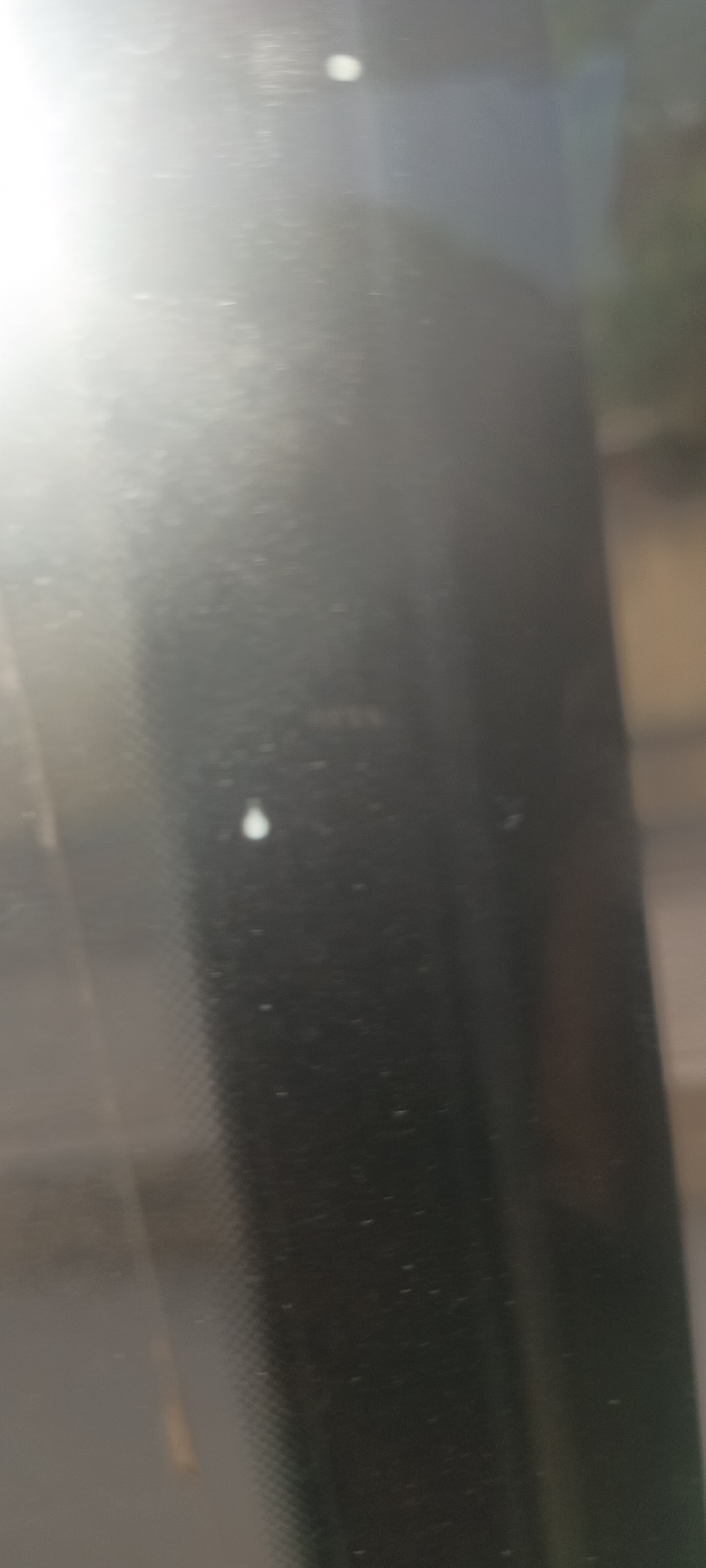 The media file displays a close-up, out-of-focus view through a glass surface, likely a window or windshield, that is covered in numerous small particles or dust. A strong, bright light source, presumably the sun, dominates the upper left portion of the image, causing significant glare and lens flare.
No people are visible in the image. The primary objects are the transparent glass surface and the various dust particles or small debris adhering to it. Two distinct, larger white shapes are visible: one near the top center and another more prominent, teardrop-shaped one in the lower-left quadrant. These could be water droplets, smudges, or larger pieces of debris.
The background seen through the glass is indistinct and blurry, appearing as dark, muted tones with faint suggestions of structures or natural elements, particularly a light brown or tan area towards the right edge.
There is no specific activity taking place, rather the image captures a static view through the obscured glass. The bright light source indicates that the photo was taken during the daytime. No text is visible within the frame. The location context is Jos, Nigeria, though this is not explicitly visible in the image itself.