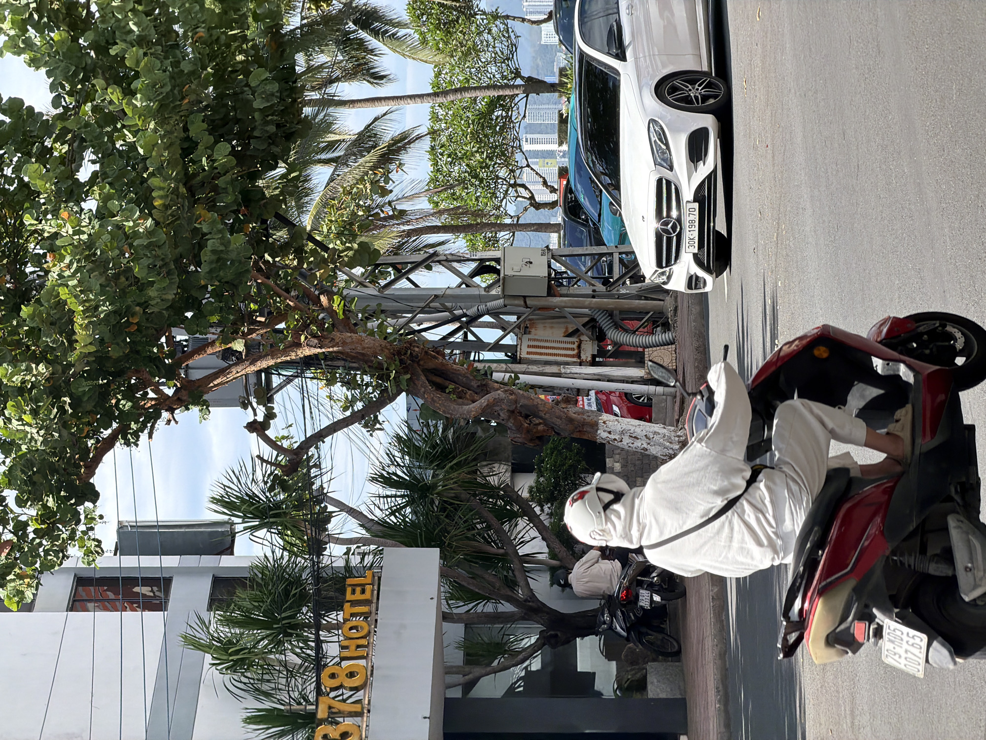 This bright, sunny photograph captures a street scene in what appears to be a coastal urban area, consistent with Nha Trang, Vietnam.
In the immediate foreground on the right, a person is seated on a red and black scooter. They are dressed in all white, including a long-sleeved shirt, trousers, a white helmet, and a face covering. The scooter's license plate, "79-M05 007.65," is clearly visible. Behind this scooter, another person is partially visible on a black scooter.
Parallel to the street on the left, a white Mercedes-Benz sedan is parked. Its license plate, "30K-198.70," is readable. Lush, green trees with rounded leaves and several palm trees line the street, creating a tropical atmosphere. Interspersed among the trees are utility poles and a metal structure with visible wiring and an electrical box.
A multi-story building with a modern white facade and dark windows occupies the midground on the left. A prominent golden sign spells out "378 HOTEL" across its lower section. In the background, beyond the trees and vehicles, a skyline of numerous high-rise buildings is visible, suggesting a dense urban environment beside what appears to be a coastline or body of water, with distant hills under a clear blue sky. The scene is bathed in bright daylight, indicating good weather.