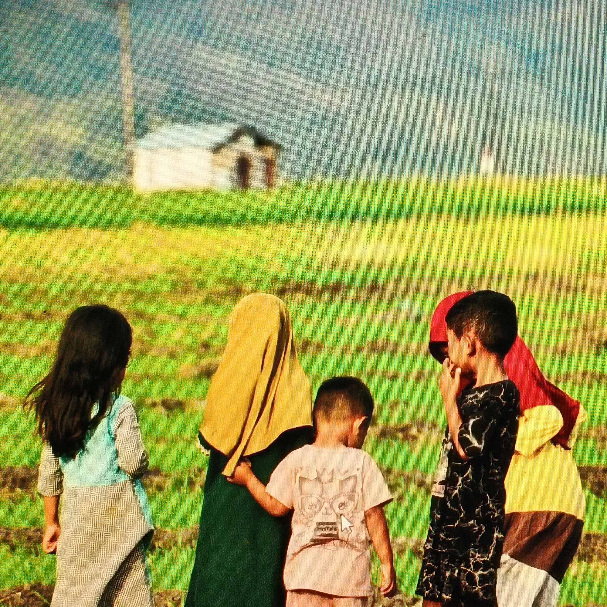 In a rural landscape near Ende, Indonesia, five children stand in a field of green, possibly rice paddies. A small, light-colored building with a dark roof sits in the background, juxtaposed against a misty, blue-gray hillside. The children are diverse in age and attire.

On the left, a young girl with long, dark hair faces away from the camera, wearing a light blue top over a patterned dress. Next to her, a person in a mustard-yellow headscarf and a dark green robe stands with their back to the viewer. To their right, a young boy in a light pink t-shirt with a graphic print and light-colored shorts looks towards the right side of the frame, his hand near his mouth. Further to the right, another child, seen from the back, wears a black and white patterned dress and a red head covering. Beside this child, another individual is partially visible, wearing a yellow top and dark pants. The children appear to be observing something in the distance, their attention drawn away from the viewer. The overall impression is one of a quiet, pastoral scene.