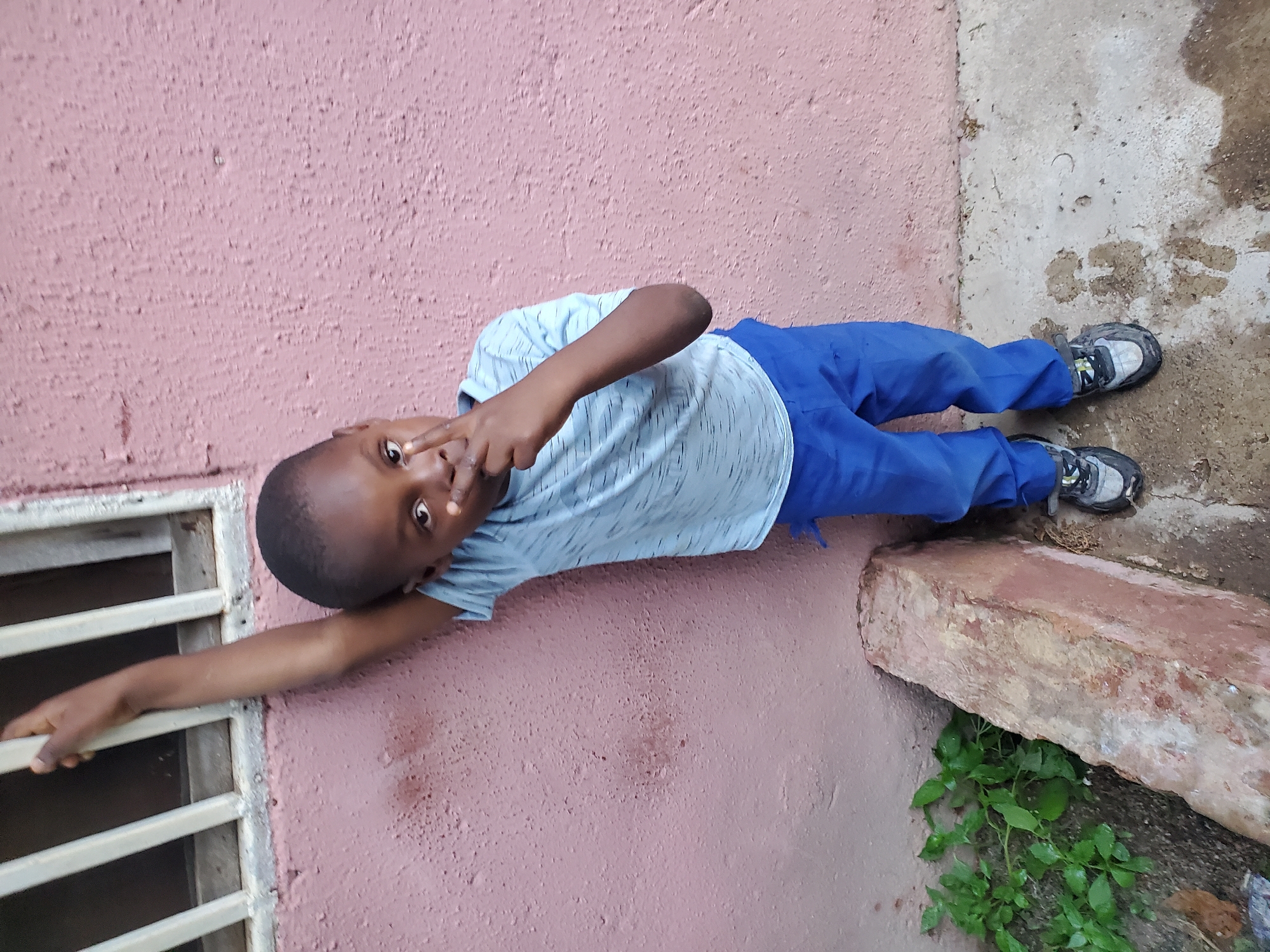 In Jos, Nigeria, a young boy stands leaning against a pink, textured wall. His left arm is extended upwards, hand resting on a white-barred window frame. He wears a light blue t-shirt with a subtle stripe pattern and vibrant blue pants. His right hand is raised, fingers forming a peace sign near his face. The ground beneath him is concrete, marked with some wet patches and debris, with a small patch of green plants growing near the wall, adjacent to a reddish, weathered brick. The scene appears to be outdoors, possibly in a residential area.
