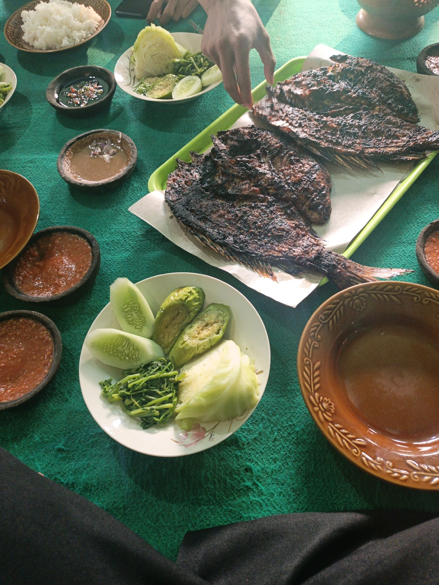 An overhead shot captures a close-up view of a table laden with a traditional Indonesian meal in Bago Krajan Dua, Indonesia. The table is covered with a textured green cloth. In the center, two whole grilled fish, charred to a dark brown, are presented on a green plastic tray lined with white paper. To the left of the fish, a white plate holds an assortment of fresh vegetables, including sliced cucumber, what appear to be steamed or boiled green beans, and wedges of boiled cabbage. Two small, dark clay bowls filled with a vibrant red chili paste (sambal) sit to the left of the vegetable plate, alongside another small bowl containing a lighter brown dipping sauce with chopped red onions. Further left, a rustic brown bowl holds a portion of white rice. In the top right, a decorative, light brown ceramic bowl with intricate carvings is visible. A human hand with slender fingers is seen reaching towards the grilled fish, suggesting the meal is about to be served or shared. The overall scene depicts a communal dining experience, highlighting the fresh ingredients and preparation methods characteristic of Indonesian cuisine.