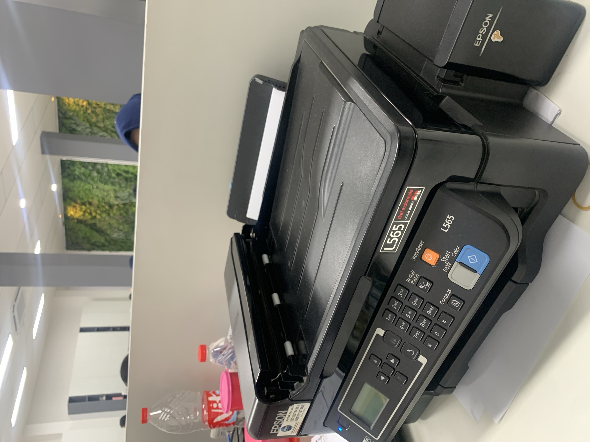 The image shows a black Epson L565 printer on a white desk. The printer has a control panel with buttons and a small LCD screen. There are also two plastic bottles of water and a pink container on the desk. In the background, there is a wall with a green plant installation and office cubicles. The scene appears to be in an office environment. There is no visible indication of the specific location in Palembang, Indonesia.