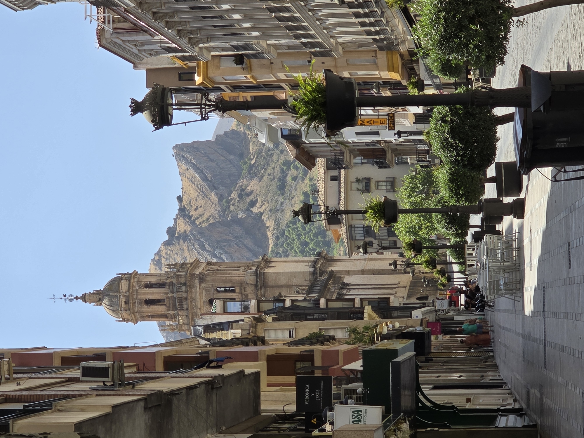 The image depicts a street scene in Jaén, Spain, dominated by architecture and natural elements. A prominent bell tower, likely part of a church, stands tall in the background, characterized by its classical design, multiple levels, and ornate dome. The tower is framed by the facades of multi-story buildings lining a paved pedestrian walkway. These buildings exhibit a mix of architectural styles, with balconies, arched windows, and various colored stonework.
On the street level, shops are visible, indicated by signage. A prominent sign reads "VERÓNICA Y JESÚS DISTRIBUIDOR OFICIAL DE MOBILIARIO." Another sign displays "ASA". Outdoor seating areas with white tables and chairs are set up along the walkway, suggesting a commercial area with cafes or restaurants. Several lampposts with decorative elements are positioned along the street, some with potted plants.
In the middle ground, a large rock formation or mountain juts out, creating a dramatic backdrop to the urban environment. Lush green trees are planted in large pots along the walkway, adding a touch of nature to the scene.
A few people are present. In the distance, two individuals are walking down the street. Closer to the foreground, a group of people are seated at one of the outdoor tables. The sky is clear and blue, suggesting a sunny day. The paving of the walkway is made of interlocking stones with darker border details. The overall impression is that of a vibrant street in a historic Spanish city, with a strong contrast between the man-made structures and the natural landscape.