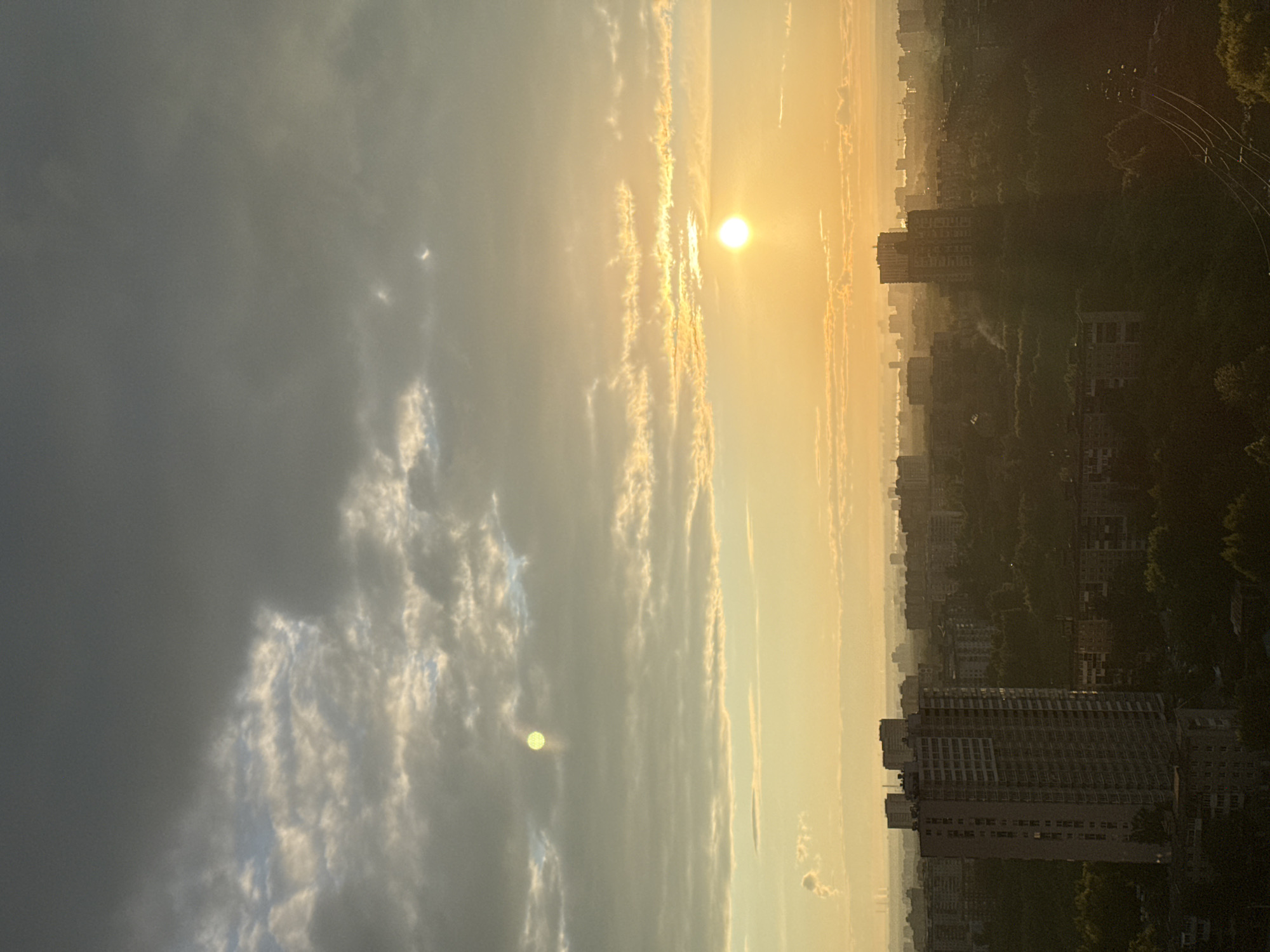 The image shows a sunrise or sunset over a city, likely Perm, Russia. The sun is visible through the clouds, casting a golden light over the scene. In the foreground, there are buildings and trees. There are power lines in the far right corner. There are wispy clouds to the left of the sun.