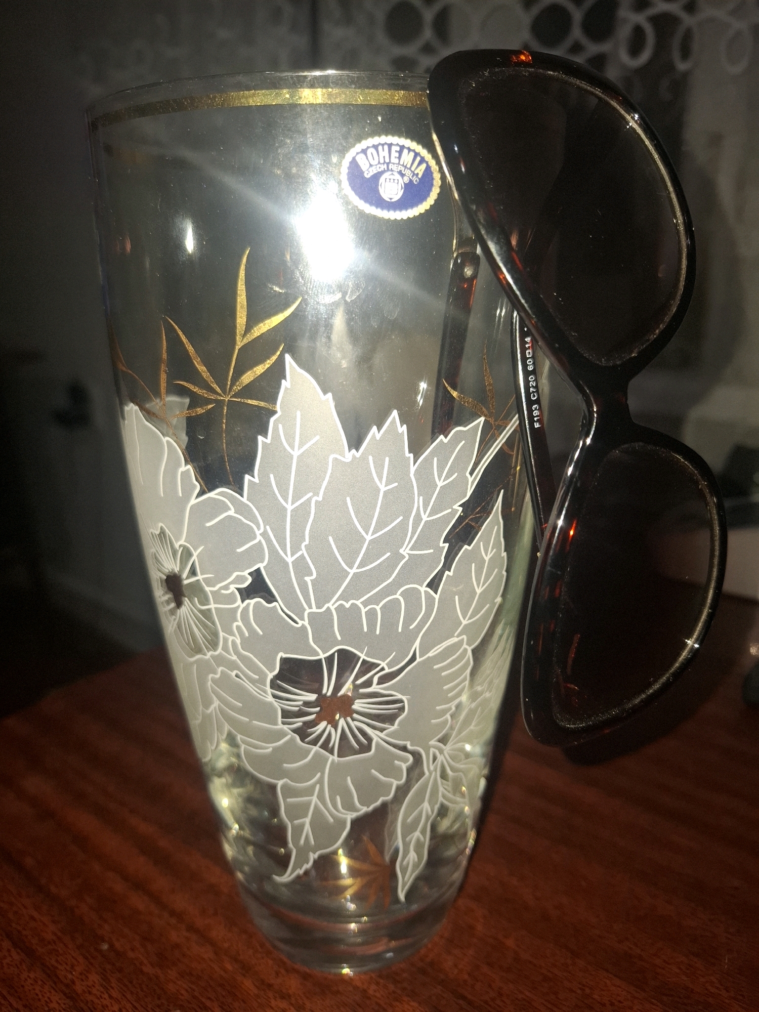 The image shows a clear glass vase with a gold rim and white floral designs. A pair of dark-rimmed sunglasses with dark lenses is resting on the vase. The vase has a sticker with "BOHEMIA CZECH REPUBLIC" on it. The vase is placed on a wooden surface. The background is out of focus and appears to be a domestic setting. There is no indication of the location being Almaty, Kazakhstan.