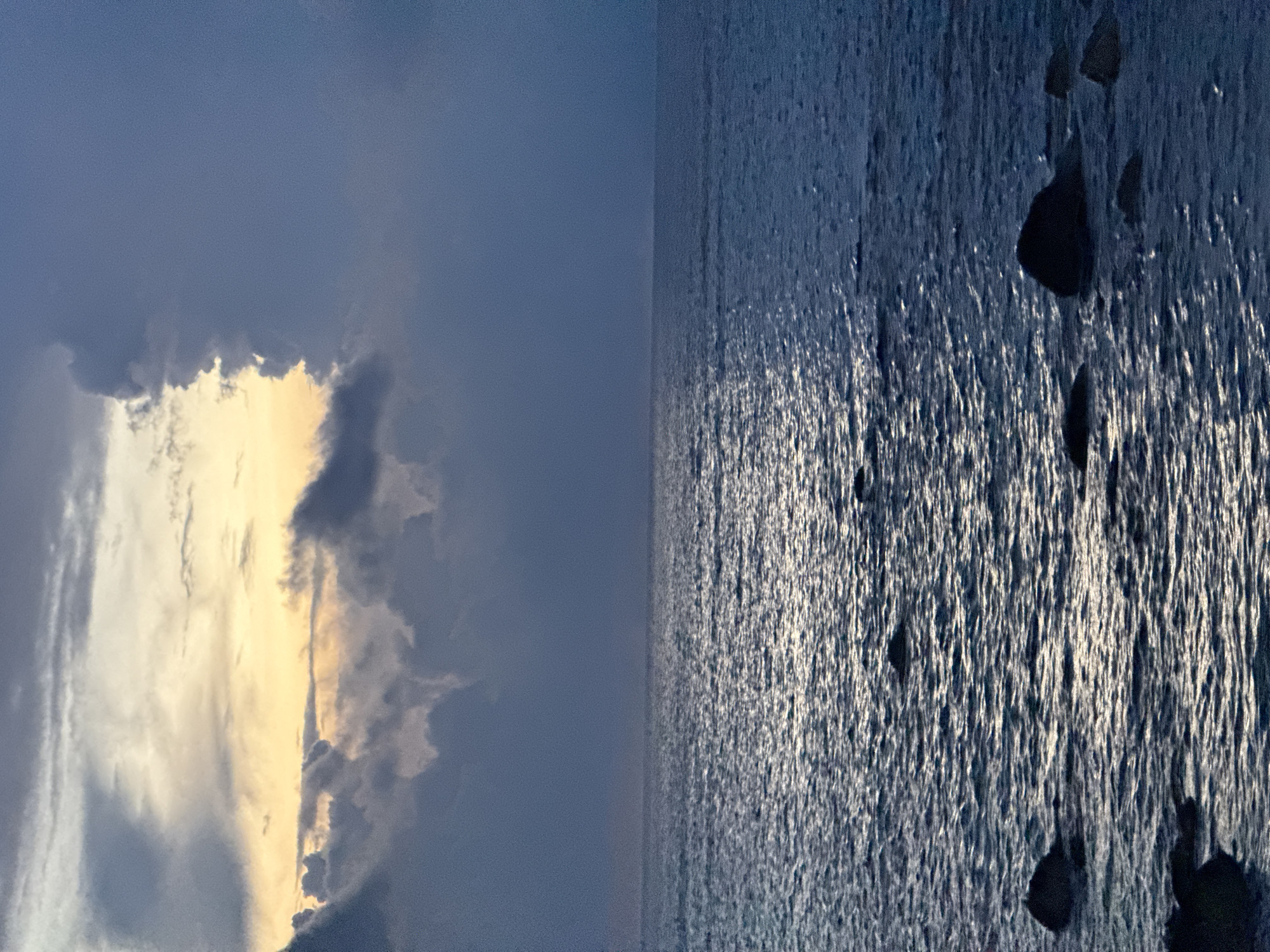 The image depicts a seascape at dusk with a dramatic sky. The foreground features dark, rounded rocks partially submerged in the choppy, dark blue ocean. The water reflects the muted light from the sky, creating shimmering streaks. The sky is a mix of dark, heavy clouds and lighter, illuminated clouds, suggesting the last light of sunset or the beginning of dawn. The illuminated clouds have a warm, yellowish-orange hue peeking through. There are no people or any man-made structures visible in the scene. The overall impression is one of natural beauty and the power of the elements. It is not possible to determine the specific location within Lý Sơn Special Administrative Region from the image alone.
