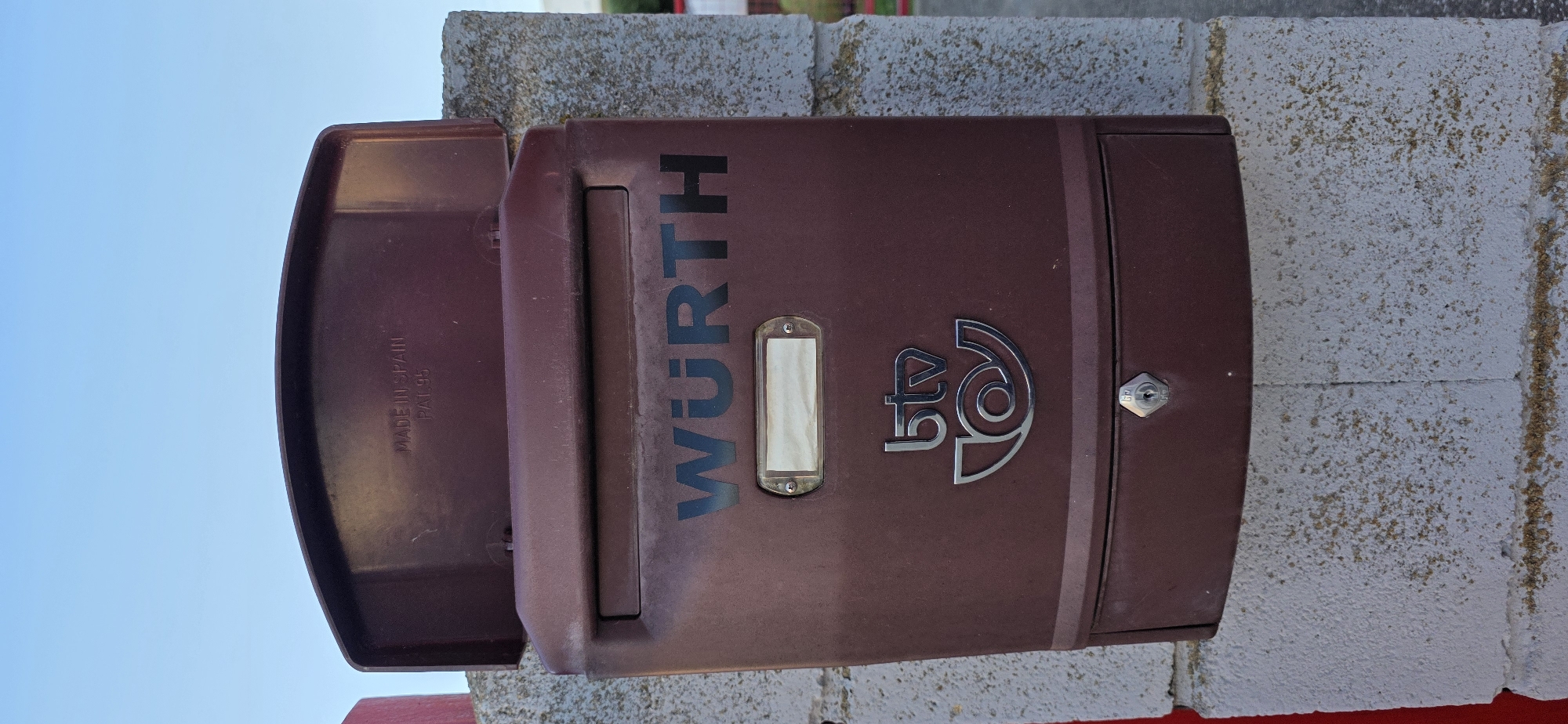 The image displays a dark reddish-brown mailbox mounted on a textured concrete block wall under a clear blue sky. The mailbox has a curved top lid that is open, revealing "MADE IN SPAIN PAT. 95" embossed on the underside. The front of the mailbox features the word "WURTH" in black lettering, with a small, empty rectangular label plate below it. Centered further down is a silver logo consisting of the letters "btv" above a stylized post horn emblem. A small silver lock is visible at the bottom of the mailbox. The concrete wall appears to be part of an outdoor structure, possibly a building exterior. The overall impression is of a utilitarian object in a somewhat weathered condition.