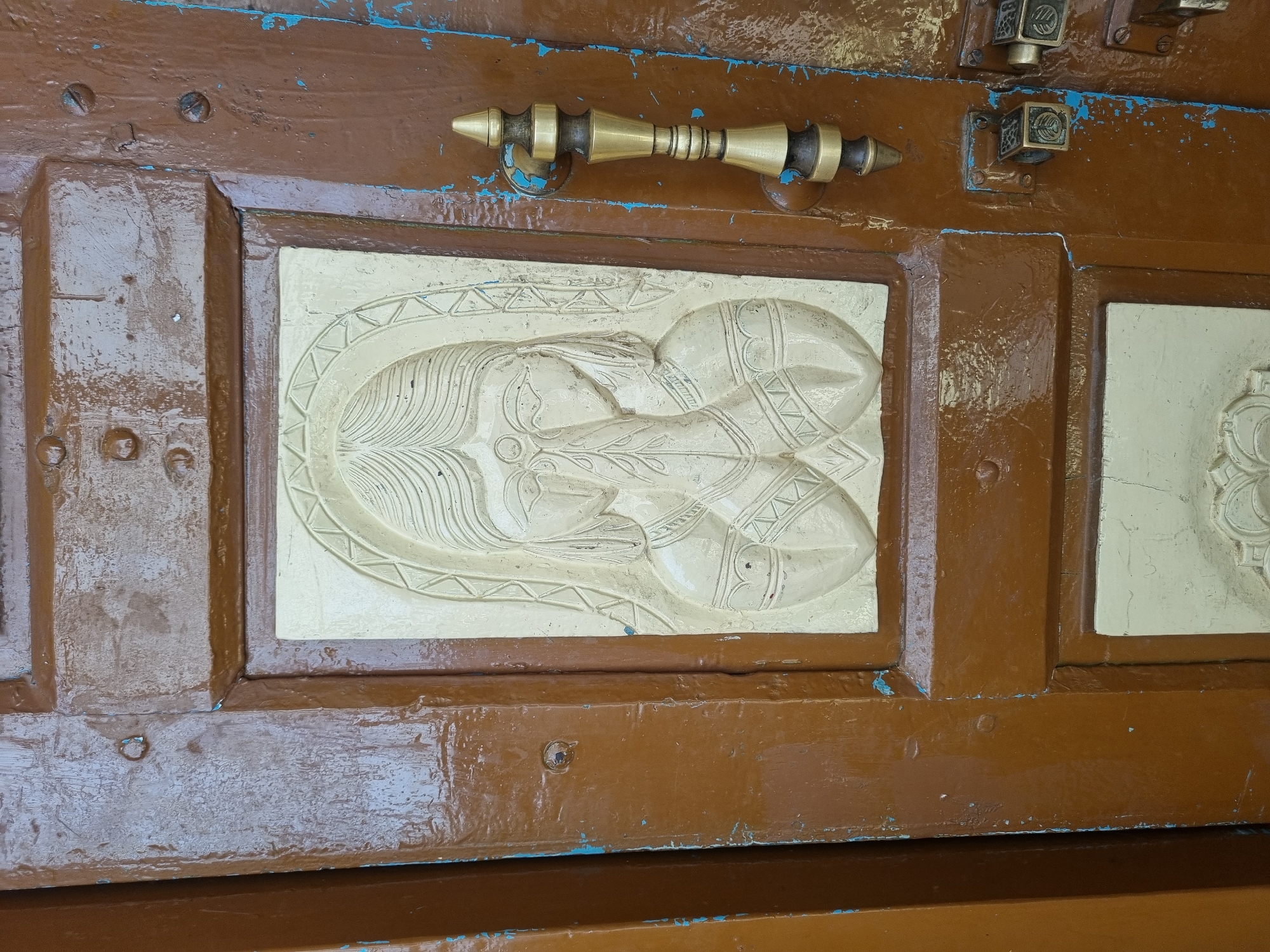 A weathered brown wooden door is depicted in Pedda Kammavaripalli, India. The door features two recessed rectangular panels with light-colored relief carvings. The upper carving portrays a stylized female figure with long hair and a head covering, her hands pressed together in a gesture resembling Anjali mudra. Her facial features are stylized, including a circular mark on her forehead, and she wears what appear to be armbands. The lower panel, partially visible, contains a circular, likely floral or geometric carving. To the right of the carvings, a vertical, ornate door handle made of brass with alternating dark bronze and gold-toned sections is affixed. Below the handle, two square, intricate metal latches or locks are visible. The brown paint on the door is chipped and peeling in multiple areas, revealing a faint blue undercoat and exposed wood beneath, indicating wear and age. Visible fasteners, including screws and nails, secure various components of the door.