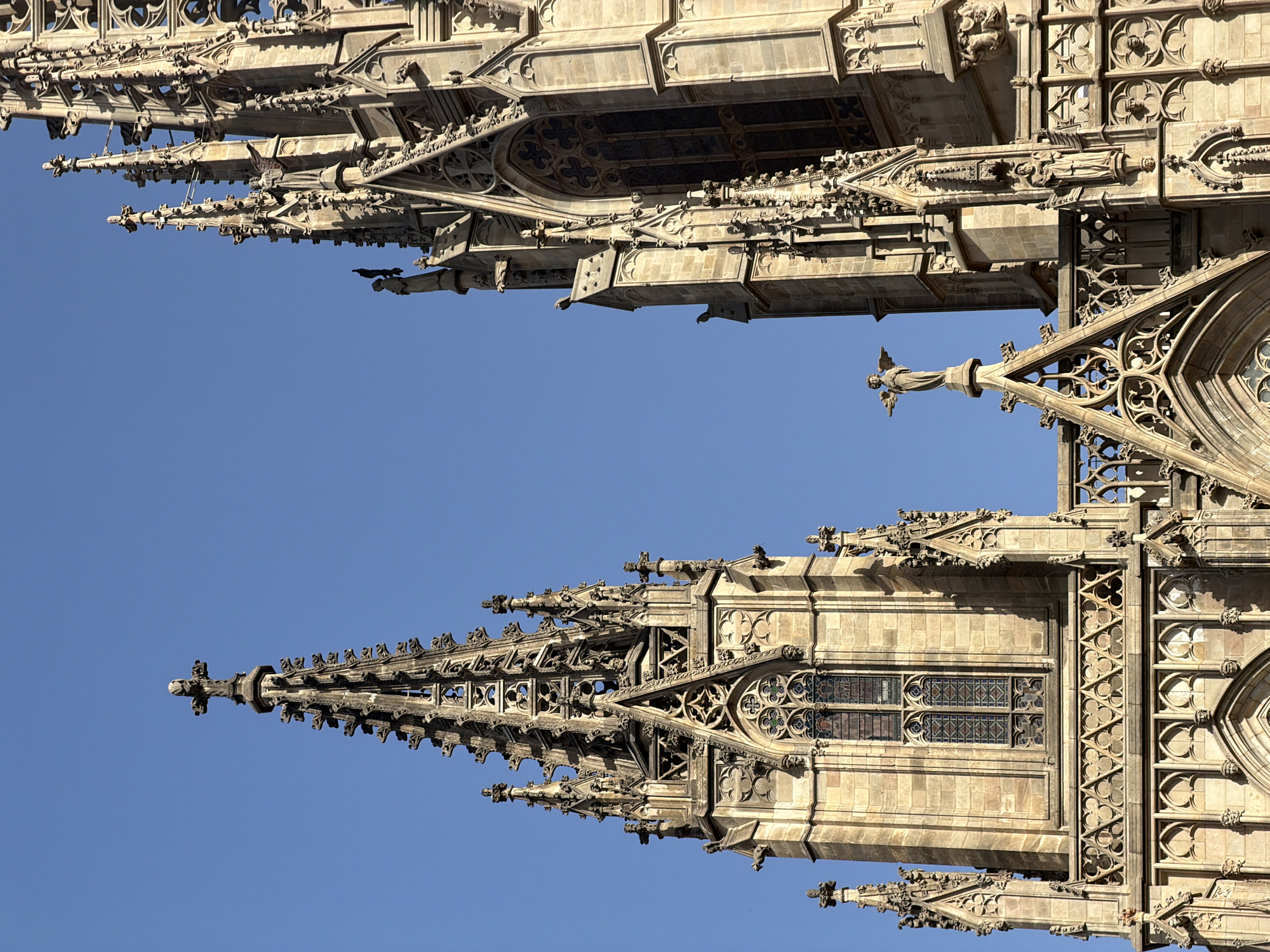The image shows the upper portions of two Gothic spires of the Barcelona Cathedral (Catedral de Barcelona) against a clear blue sky. The spires are constructed of light-colored stone and are elaborately decorated with intricate architectural details, including pinnacles, gargoyles, and tracery.
The spire on the left is taller and features a prominent cross at its apex. It has a series of tiered windows with stained glass, suggesting it is part of a bell tower or similar structure.
The spire on the right is slightly shorter and wider, with a statue of an angel standing on a pinnacle at its peak. This spire also exhibits rich Gothic ornamentation, including pointed arches, sculpted figures, and elaborate stonework.
The sunlight casts shadows that highlight the three-dimensional nature of the carvings and architectural elements. The overall scene conveys a sense of grandeur and historical significance.