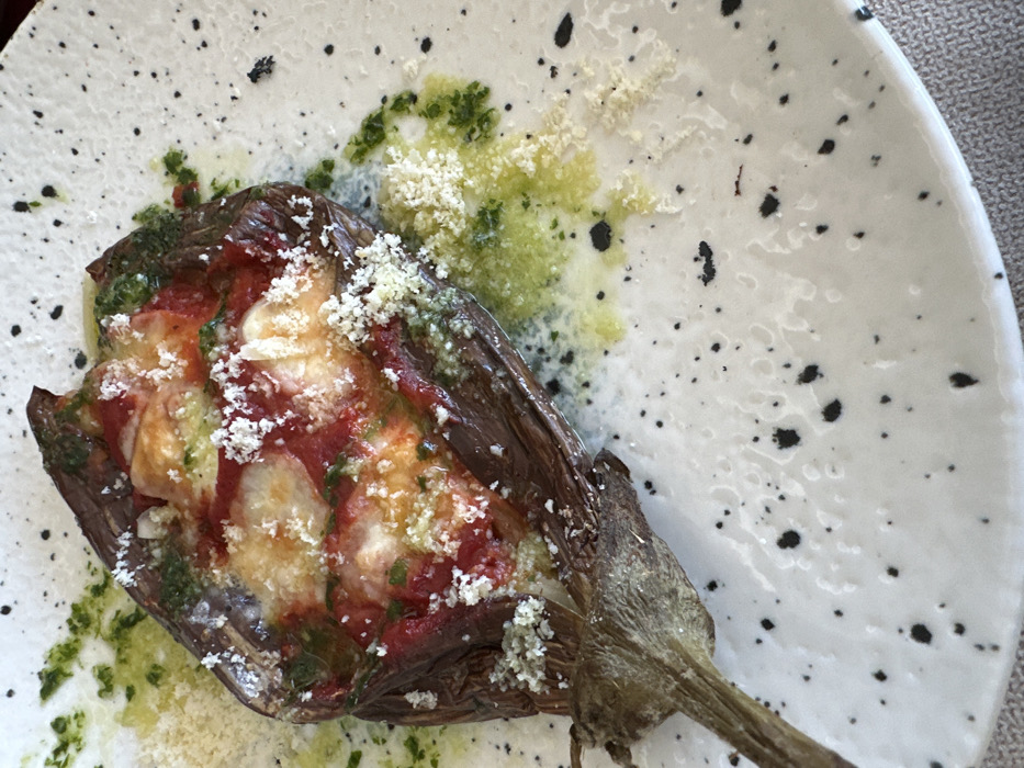 An image captured in Pancharevo, Bulgaria, displays a single baked eggplant half presented on a white ceramic plate. The eggplant, dark purplish-brown with its stem intact at the lower left, is split lengthwise and filled with a red tomato-based sauce, melted white cheese, and visible green herbs. Grated white cheese is liberally sprinkled over the top of the eggplant and onto the plate. Around the base of the eggplant, a light green, oily sauce (possibly herb oil or pesto) is drizzled, along with additional scattered grated white cheese. The plate features a textured surface with irregular black speckles. The plate rests on a light beige or off-white woven textile surface.
