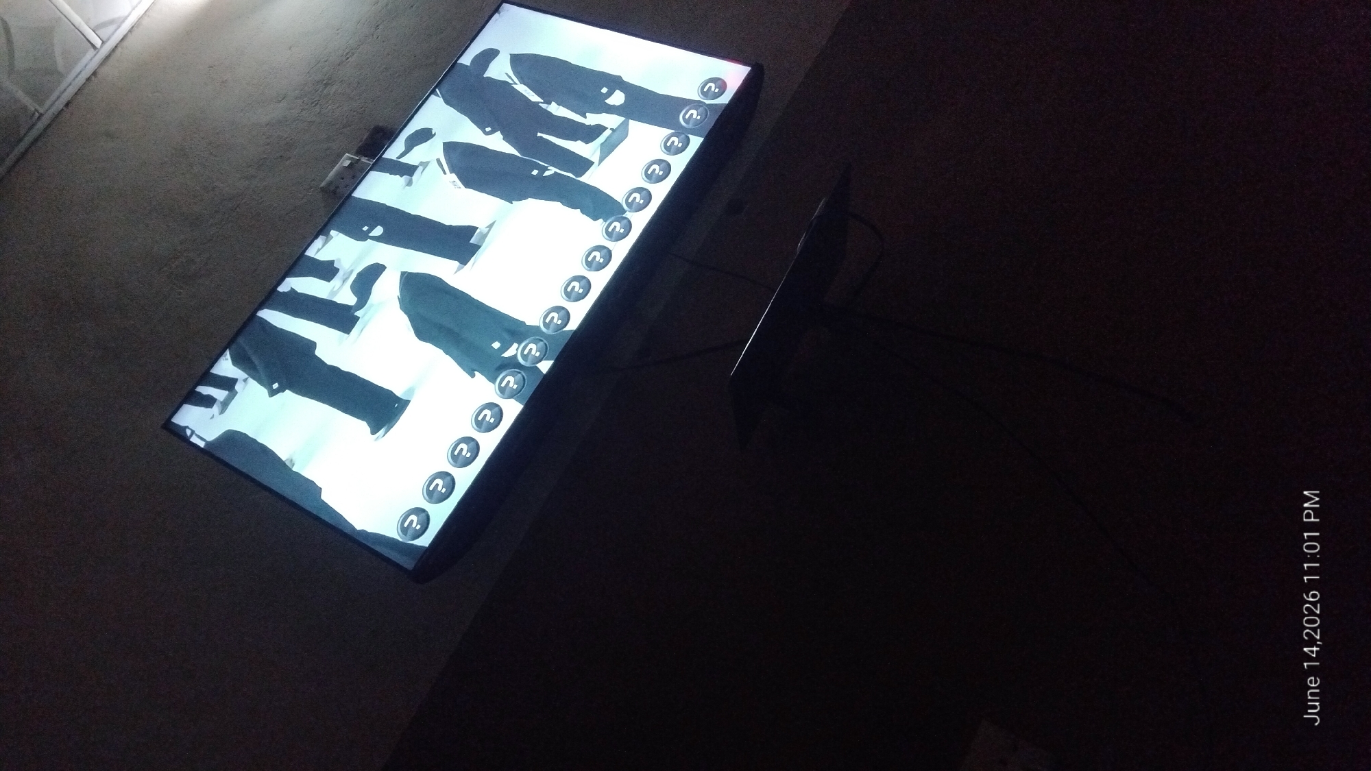 The image shows a television screen displaying a black and white scene of people wearing suits. The television is mounted on a wall in a dimly lit room. Below the television is a black shelf with some cables. The timestamp "June 14, 2026 11:01 PM" is visible in the bottom left corner of the image. The image does not provide any information about the location in the city of Howayi, Nigeria.