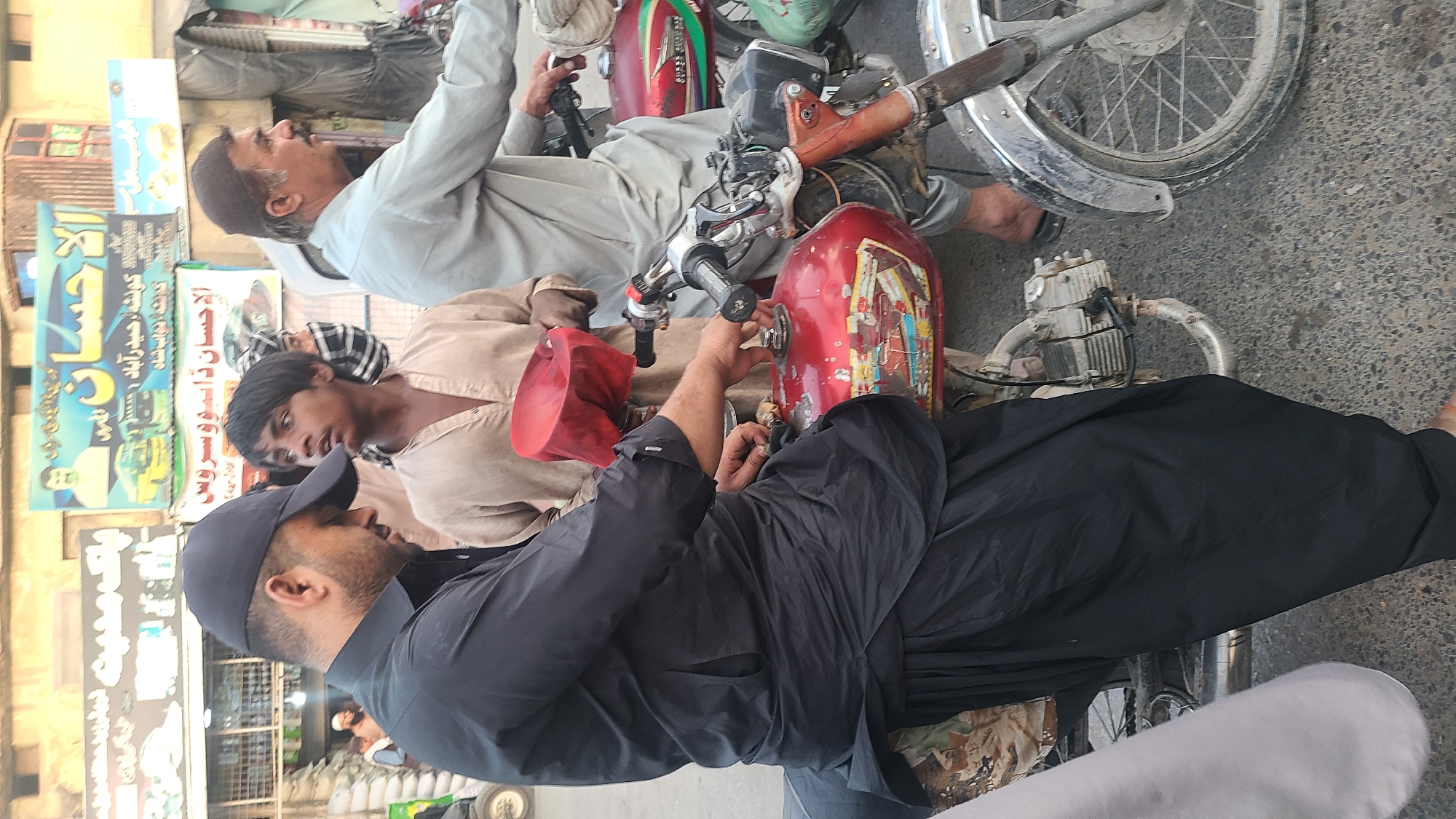 The image depicts a street scene in Quetta City Tehsil, Pakistan. Three men are present, two of them sitting on motorcycles. One man wears a light-colored shirt and a Muslim prayer cap, while a second man has a tan shirt and a black and white head covering. A third man, dressed in black attire and wearing a baseball cap, is standing near the second motorcycle. The motorcycles are parked on a paved road, and the environment includes storefronts with Arabic signage in the background. One of the motorcycles appears to be undergoing maintenance.