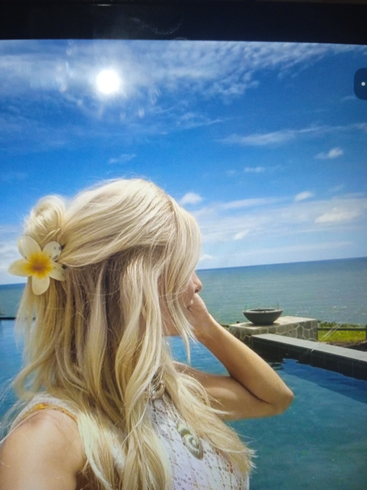 The image shows a close-up of a blonde woman with a flower in her hair, seen from the back and side, facing a body of water. She is wearing a white, patterned top. The background features a bright blue sky with some clouds and the sun. A calm ocean or large body of water is visible in the distance, with a stone structure and a dark bowl on a ledge overlooking the water. The foreground shows a bright blue swimming pool. The image does not provide any information or context related to the city of Chernivtsi, Ukraine.
