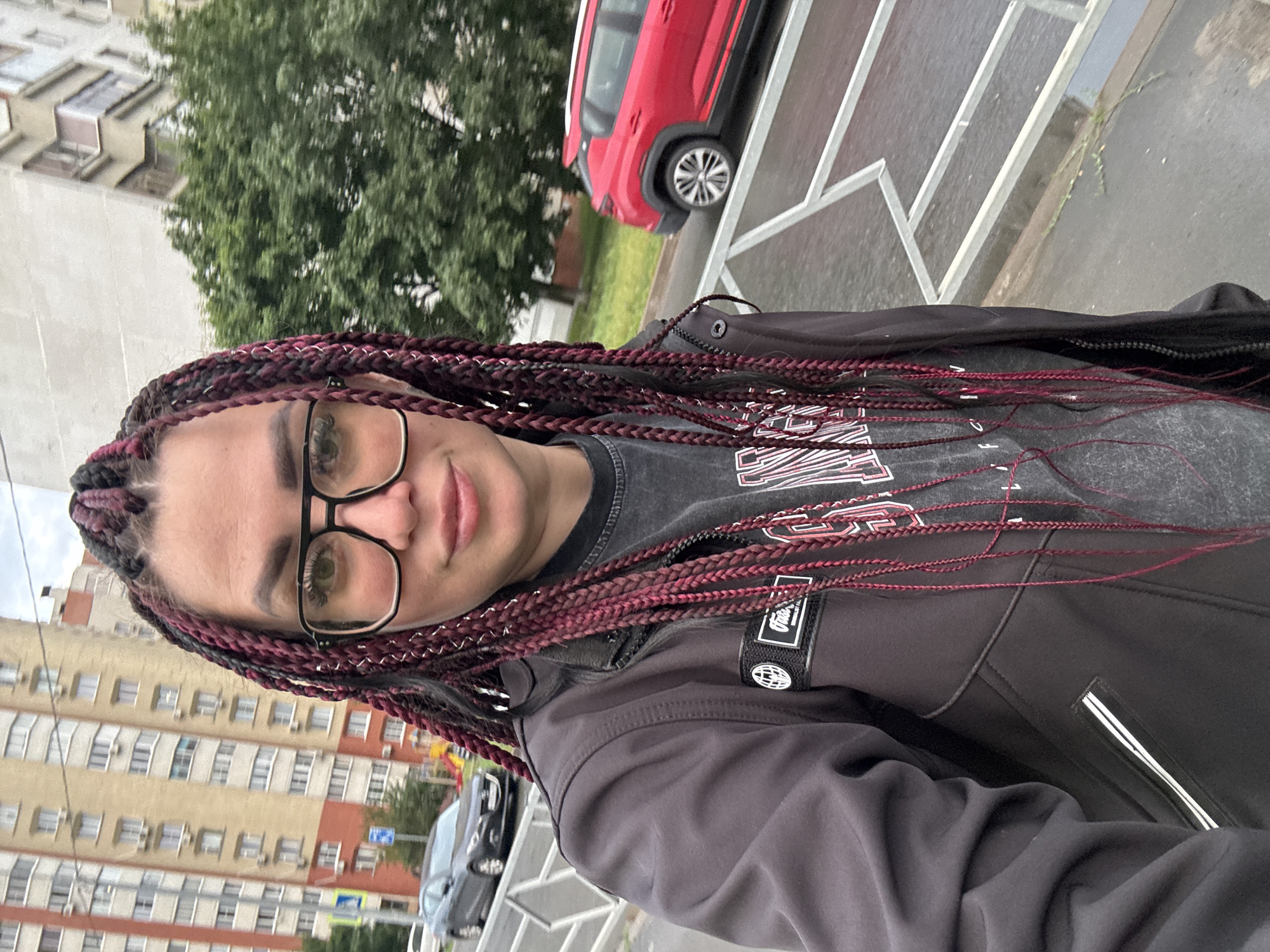 A woman with dark hair braided with maroon extensions is standing on a sidewalk. She is wearing a dark jacket over a dark t-shirt, and large-framed glasses. A red SUV is parked on the street behind her. In the background, there are multi-story apartment buildings and trees. The scene appears to be in an urban environment in Tolyatti, Russia.