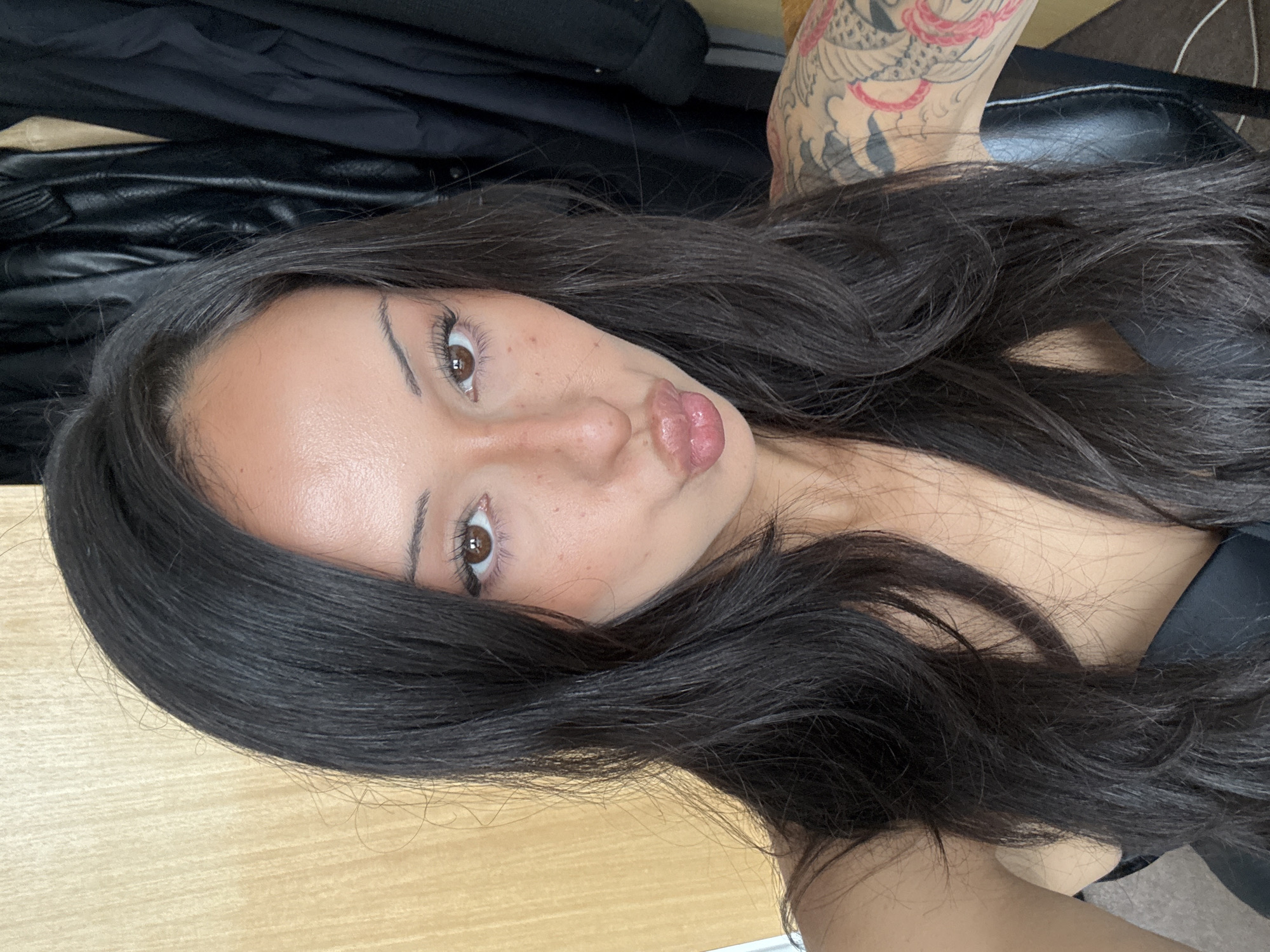 The image shows a close-up, selfie-style portrait of a woman with long, dark hair. She is wearing a black top and has visible tattoos on her left arm and shoulder. Her facial expression is one of puckering her lips. The background appears to be an indoor setting, with a wooden surface to the left and what looks like black leather clothing hanging to the right. There is no discernible information in the image that would indicate a specific location in London, United Kingdom.
