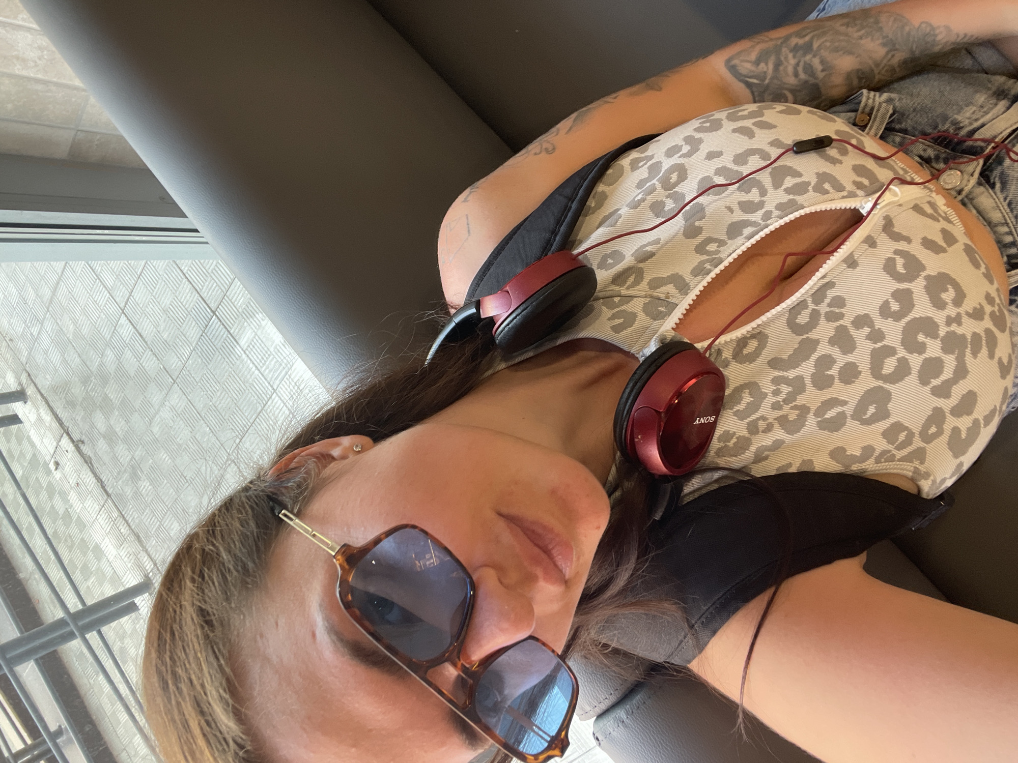 A woman with sunglasses and headphones is lying on a grey bench. The woman is wearing a patterned top, and denim shorts. Her right arm is visible, showing tattoos. The headphones are red and black and the brand is Sony. Outside the window is a sidewalk and tiles. The location is in Misaylovo, Russia.