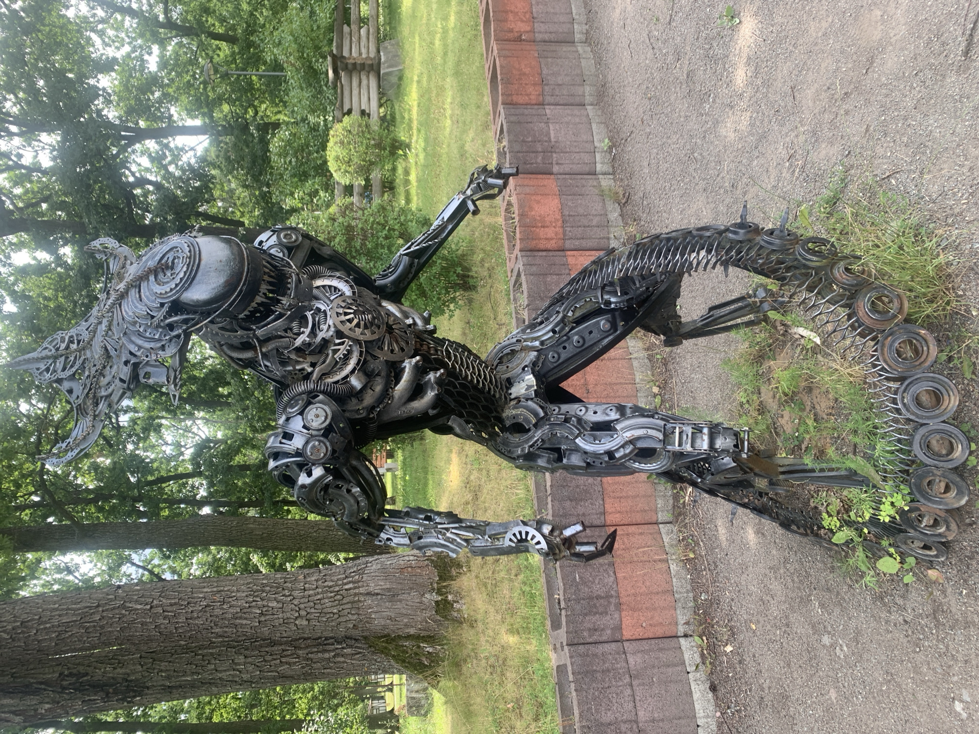 The image depicts a large, metallic sculpture of an alien creature, possibly inspired by the Xenomorph from the "Alien" franchise. The sculpture is constructed from various metal parts, including gears, pipes, and other mechanical components. It is positioned outdoors in a park-like setting with trees and foliage visible in the background. A low, brick wall runs along the middle ground. The sculpture stands on a gravelly path with patches of grass around its base. In the far background, a rustic wooden fence and some buildings can be observed, suggesting an urban or suburban park environment. This sculpture is located in Bucha, Ukraine.