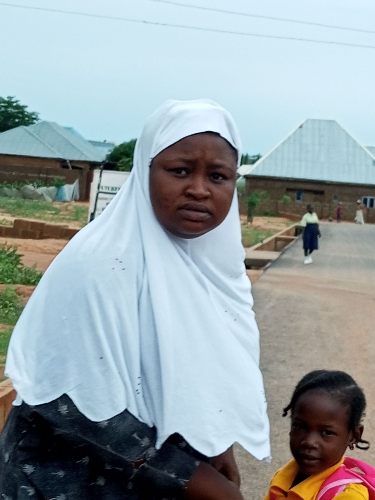 An outdoor, low-angle shot features a Black woman wearing a white hijab and a Black child with braided hair, wearing a yellow shirt and a pink backpack. The woman is positioned in the foreground, her face turned slightly to her left and her gaze directed forward. Her expression is neutral. The child is in the lower right corner, looking towards the camera with a faint smile.
In the background, several buildings with light-colored, sloped roofs are visible, suggesting a residential or institutional setting. Greenery and some construction materials are also present. A person in a dark skirt and light-colored top is walking away from the camera on a paved path. The sky is overcast and pale blue. The scene appears to be in Gombe, Nigeria.
