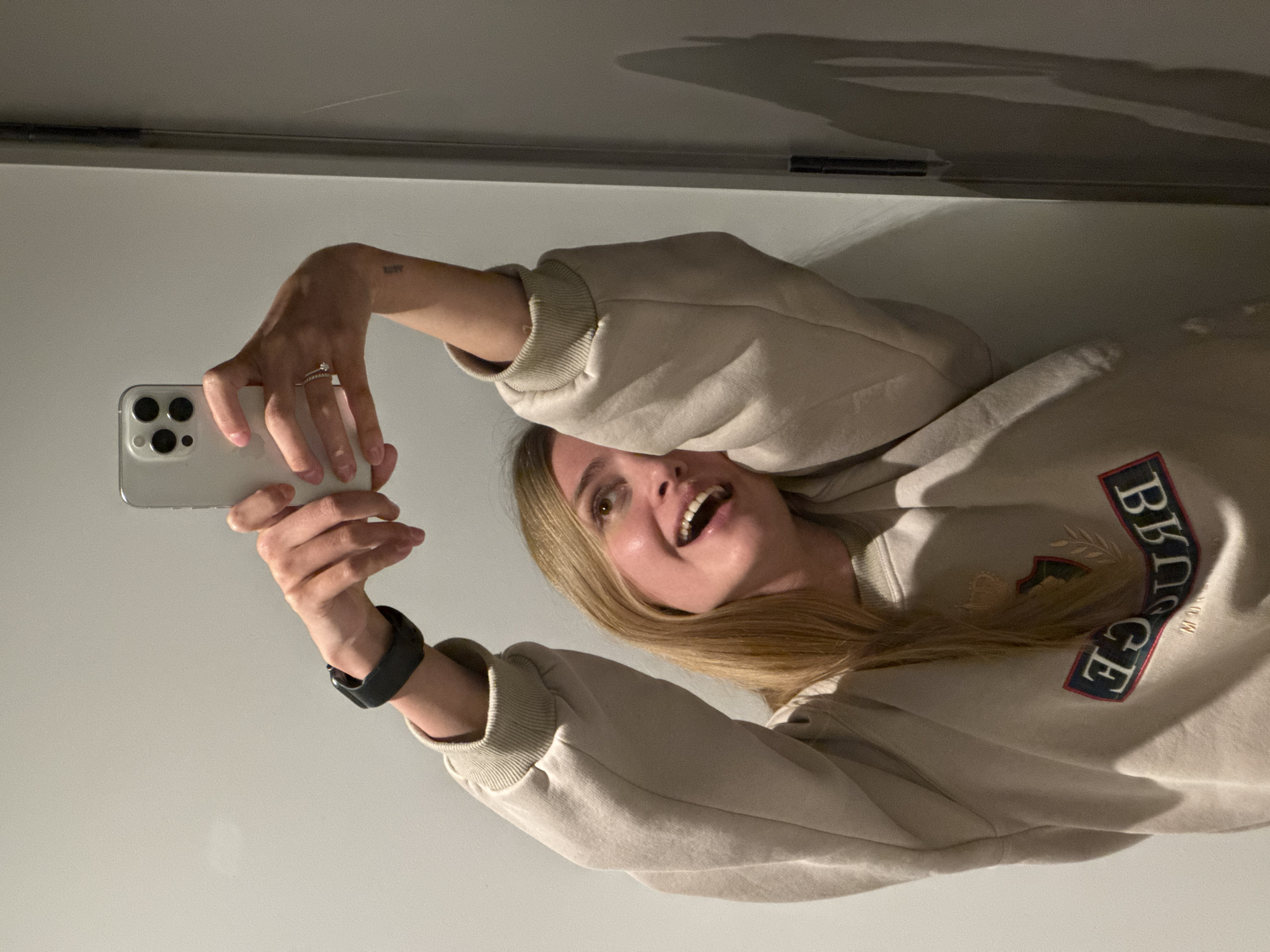 A woman is taking a selfie indoors, holding a white smartphone. She is wearing a beige sweatshirt with the word "BRIDGE" embroidered on it, and a black smartwatch on her left wrist. She also has a ring on her left hand. The background consists of a plain wall and ceiling. The location in Tbilisi, Georgia is not discernible from the image.