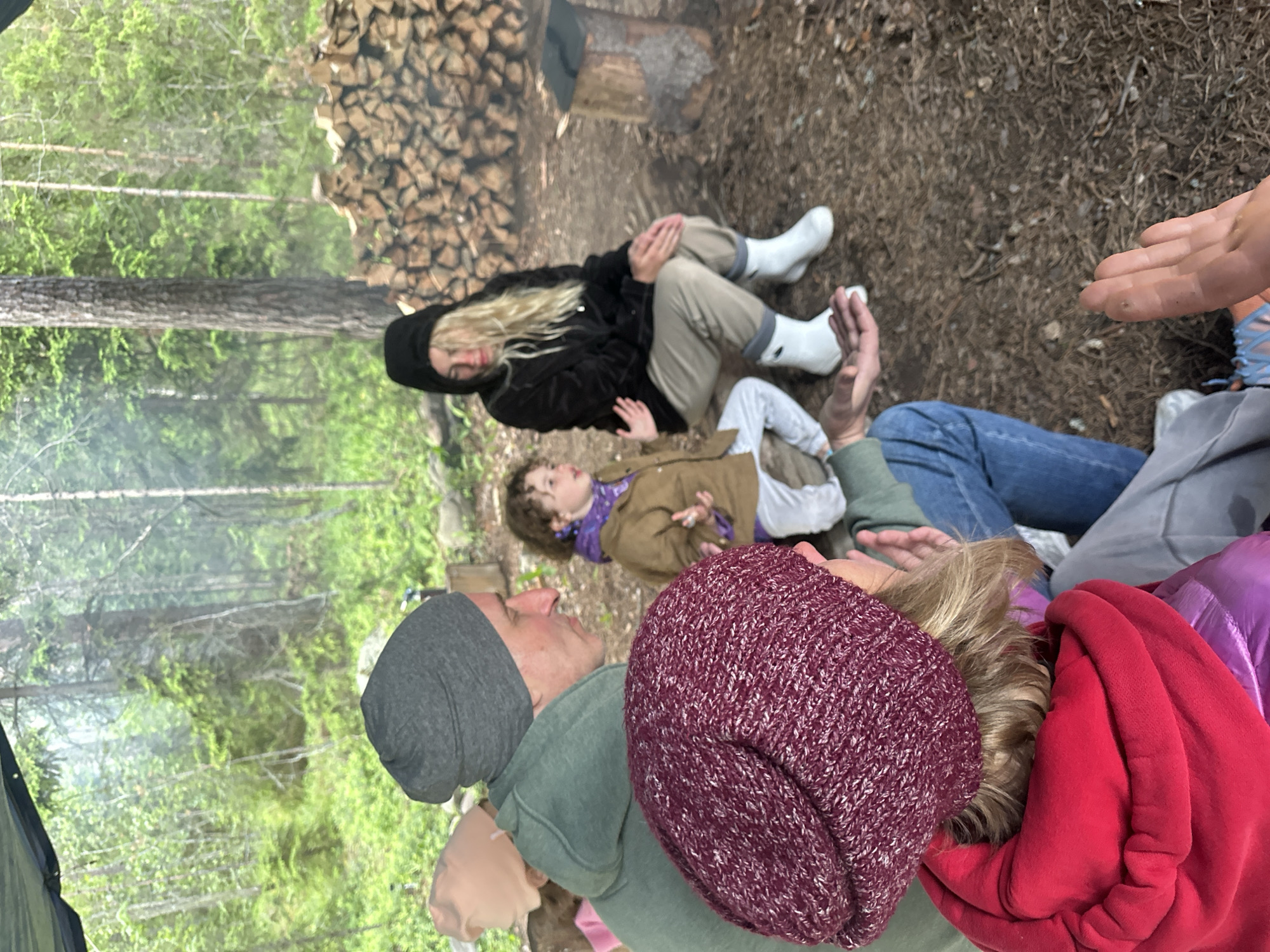 In the city of Soanlakhti, Russia, a group of four individuals, two adults and two children, are gathered around a fire pit under a canopy in a forest. The scene appears to be a casual outdoor gathering. One adult, a woman with blonde hair and wearing a black hooded jacket and light-colored pants, is seated with her hands clasped. Beside her, a young child in a brown jacket and purple scarf looks to their right. In the foreground, another adult, wearing a dark red knitted hat and a red hooded jacket, is partially visible, their hand outstretched as if gesturing or interacting with someone off-camera. Next to this individual, a man wearing a grey beanie and a green hooded sweatshirt is also visible, looking to his right. The environment is natural, with trees and a pile of firewood in the background. The ground is covered with pine needles and debris. A small campfire or fire pit is visible to the right.