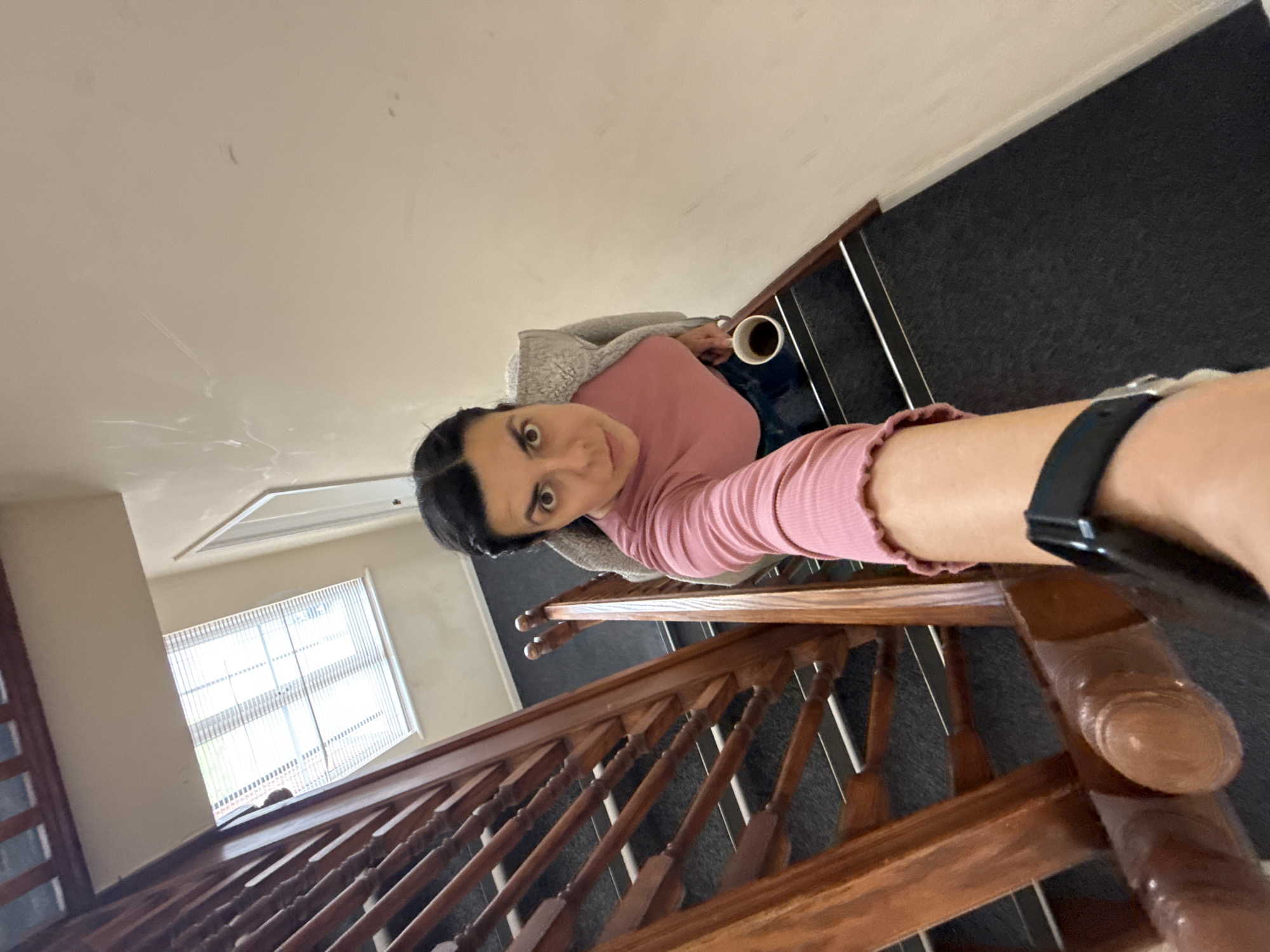 A high-angle indoor shot features a person on a staircase. The person, a female with dark hair pulled back, is wearing a pink long-sleeved shirt and a grey hooded jacket. She is holding a cup in her left hand. The staircase is wooden with a dark carpet on the steps. A black smartwatch is visible on her wrist closest to the camera. A window with blinds is visible in the background, along with a section of the ceiling. The location is plausibly a residential building in Great Yarmouth, United Kingdom, given the typical architectural features, although this cannot be confirmed from the image alone.