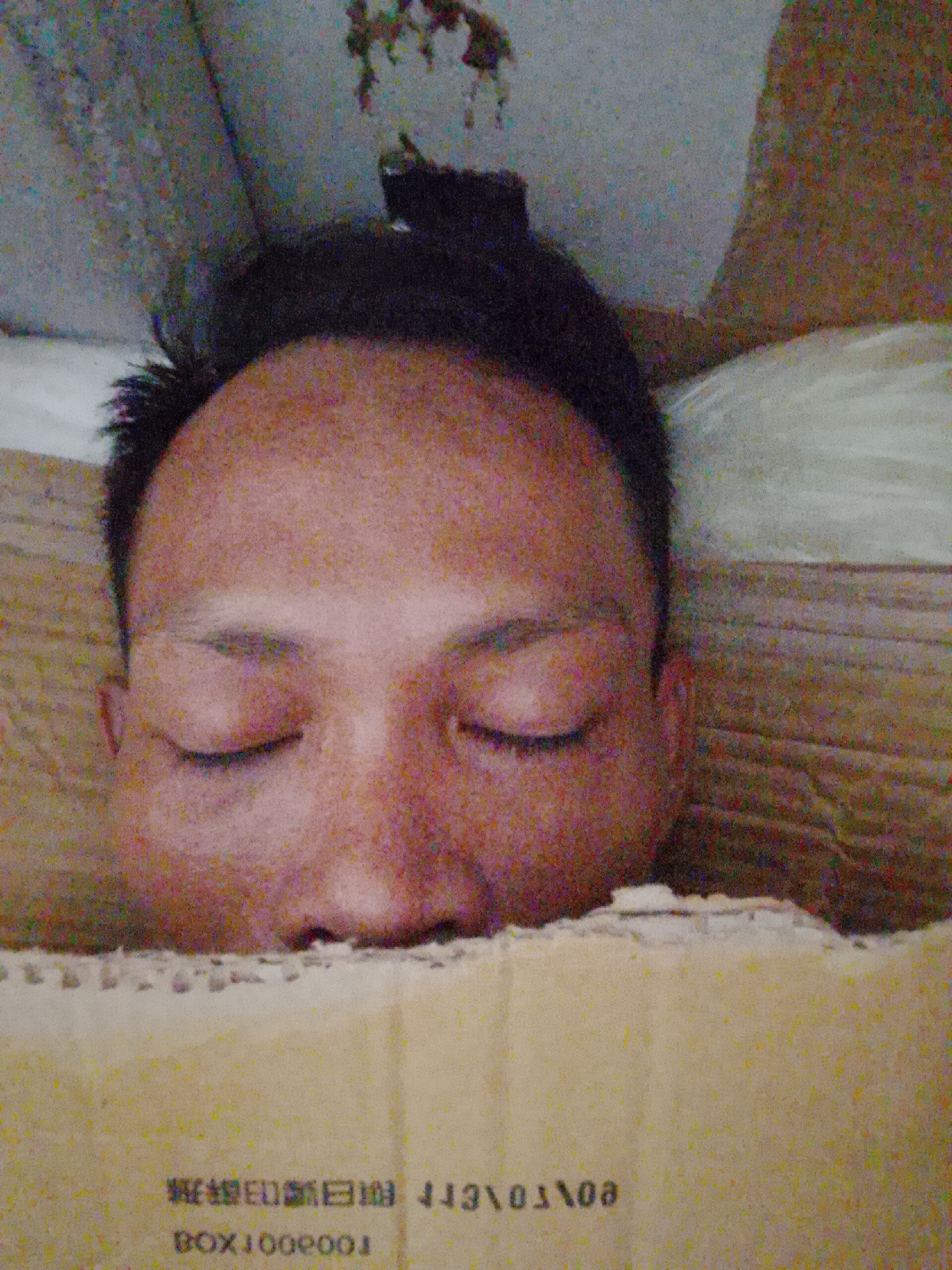 A medium, eye-level shot shows a person with dark hair and closed eyes partially obscured by a cardboard box. The person's face is visible from the forehead to the top of the nose. The cardboard box has text printed on it. The background includes a wall or other structure. The image's location is Bukit Ketok, Indonesia, although nothing in the image confirms this.