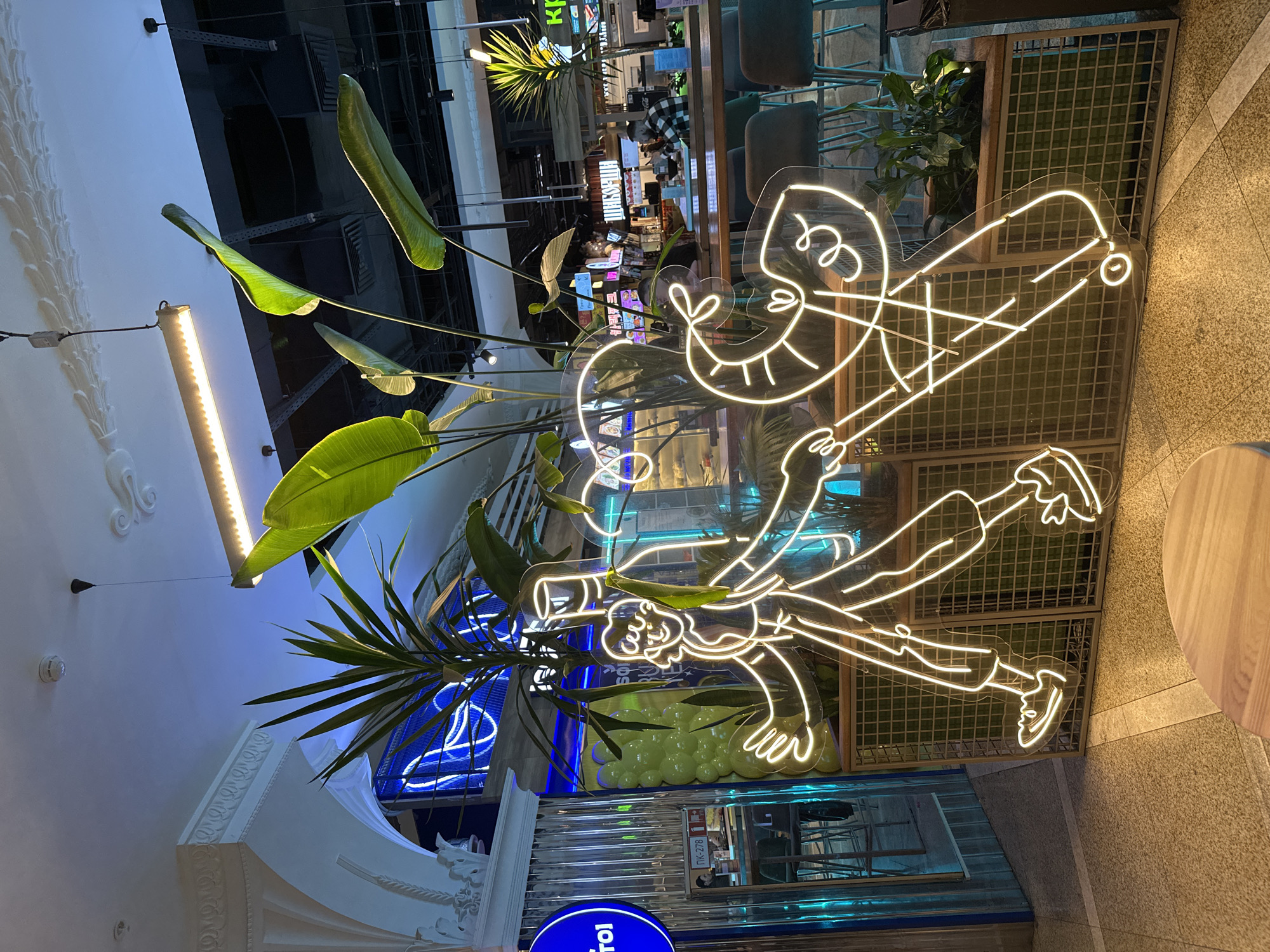The image captures an indoor scene, possibly a restaurant or cafe, in Yekaterinburg, Russia. A neon sign depicting a stylized man with an exaggerated grin and bowler hat stands out. It features simple lines, suggestive of movement or dance. Lush green plants are interspersed around the sign, adding a touch of nature to the interior. The background includes glimpses of the establishment's interior, showing seating areas, counters, and various decorative elements like lights and geometric patterns. The overall atmosphere is modern and vibrant.