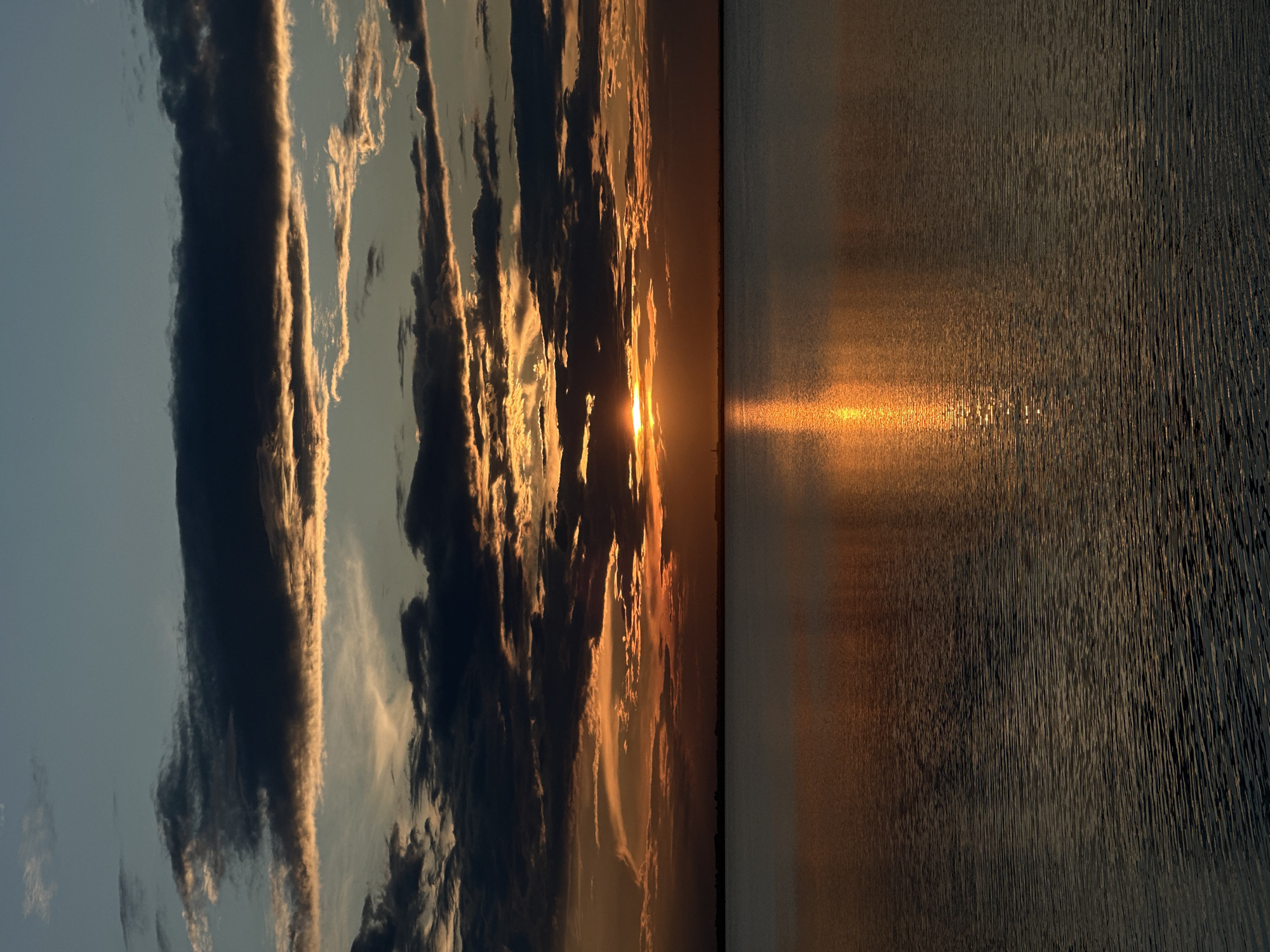 The image captures a dramatic sunset over a body of water. The sky is filled with a mixture of dark, brooding clouds and streaks of vibrant orange and yellow as the sun sets behind them. The sun itself is a bright, concentrated orb peeking through a gap in the clouds. The light from the setting sun is reflected intensely on the surface of the water, creating a shimmering, golden path. The water itself appears calm, with gentle ripples. A faint, silhouetted line of land is visible on the horizon, with what appears to be a small structure or tower.
The scene is a natural landscape and does not depict any specific identifiable landmarks within Kokshetau, Kazakhstan. The image focuses on the atmospheric conditions of a sunset over water. There are no people, actions, or interactions visible in the photograph. The environment is primarily sky and water.