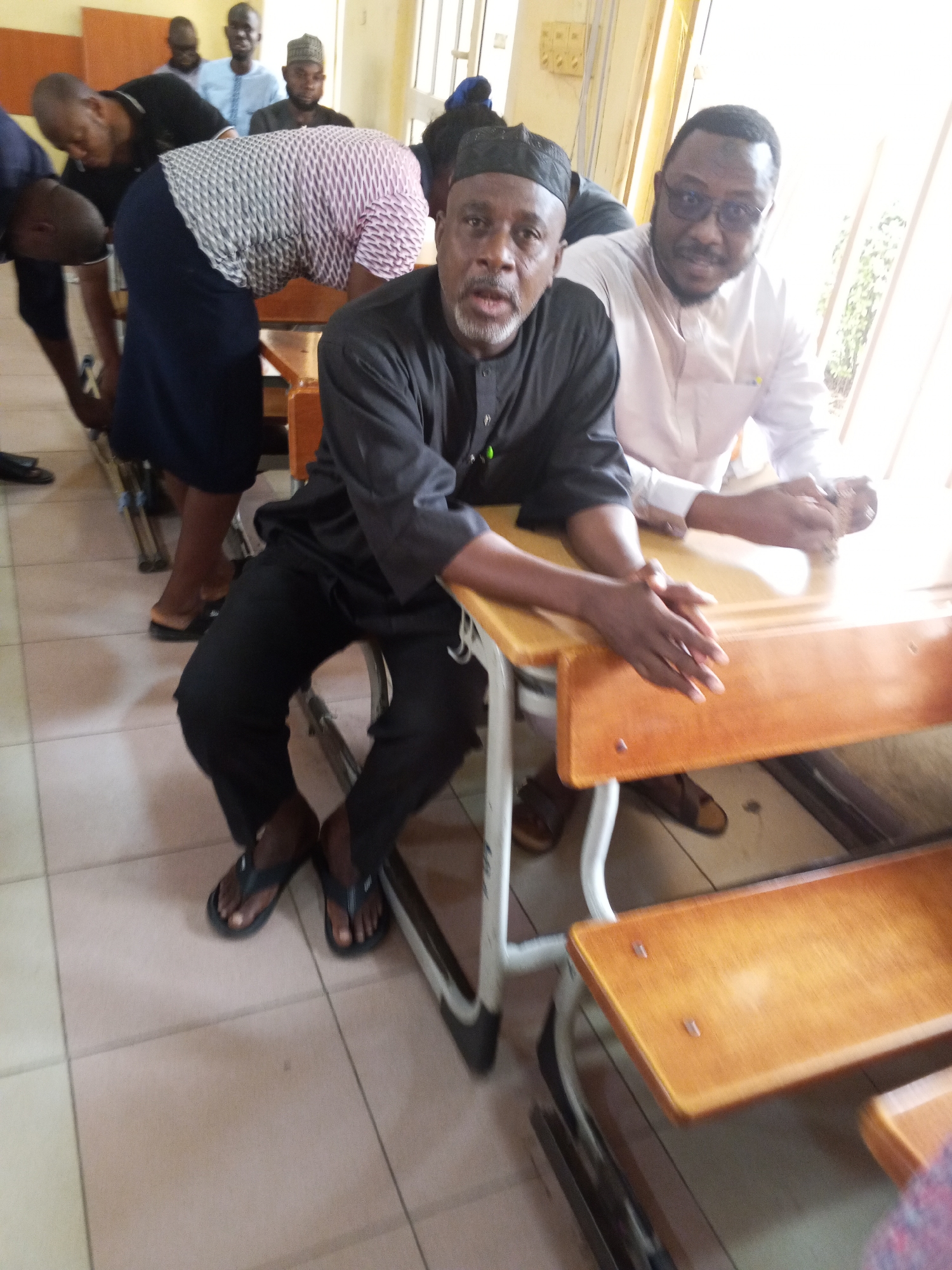 The image shows a group of men in what appears to be a classroom or meeting room setting. In the foreground, a man with a gray beard and a black cap is seated, looking towards the camera. He is wearing a black traditional outfit. To his right, another man wearing glasses and a light-colored shirt is also seated, looking to his right. Several other men are visible in the background, some standing and others seated, suggesting a gathering or a lecture. The room has a tiled floor and light-colored walls, with a window or opening visible on the right side, letting in natural light. The overall atmosphere seems formal, possibly a discussion or educational event. The men are dressed in a mix of traditional attire and casual wear. There are wooden desks with metal frames, typical of a school environment. The scene is indoors, and no information about the time of day or weather is explicitly provided. The location context is Lokoja, Nigeria.