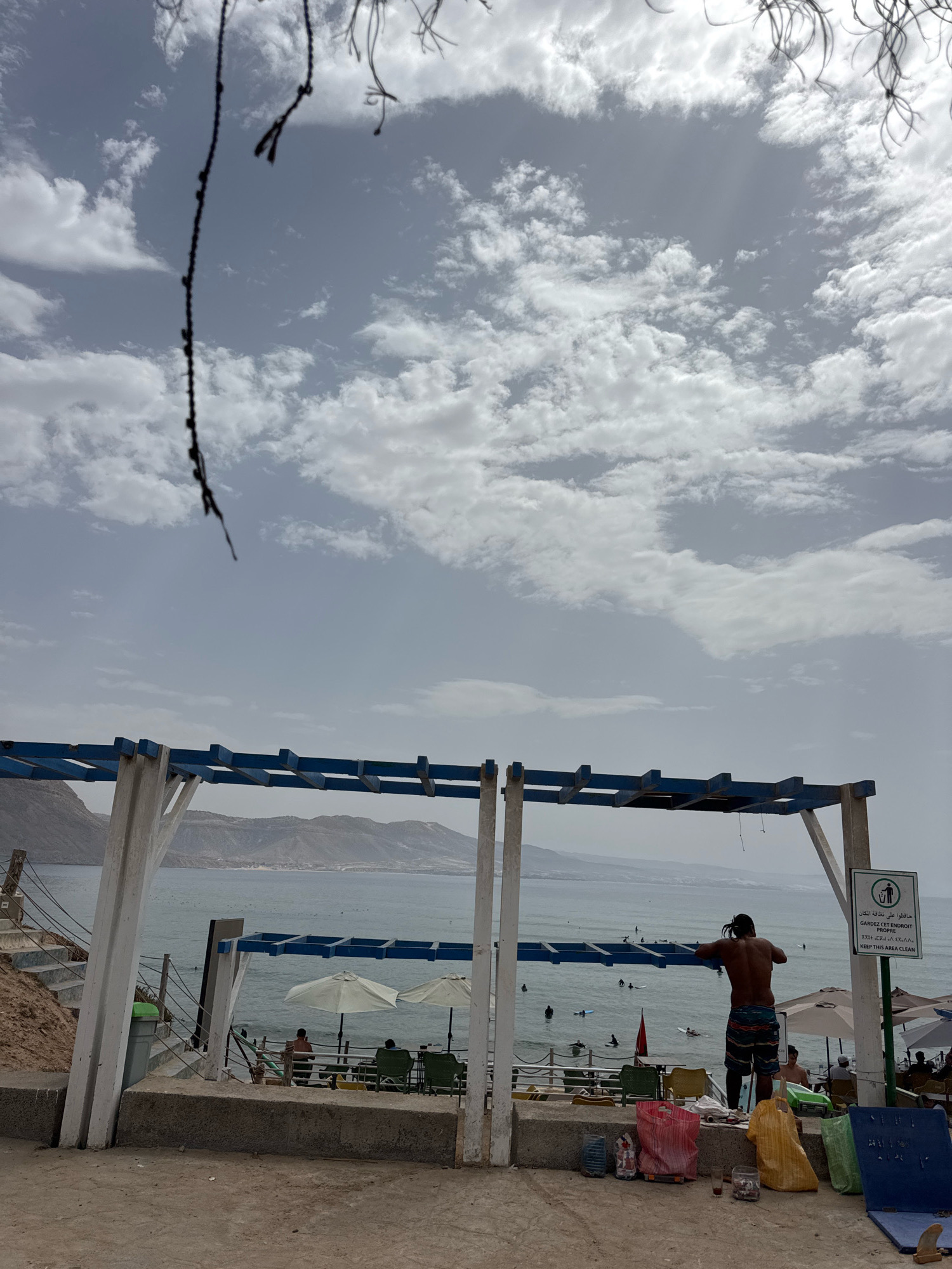 In Imsouane, Morocco, a man stands with his back to the camera, wearing patterned swim trunks. He is positioned near a large, blue-framed awning structure that extends over a beach area. In front of him, several people are visible in the ocean, some on surfboards. To the right of the man, a green sign with white lettering, bearing a recycling symbol, advises to "KEEP THIS AREA CLEAN." Below the awning, there are chairs, umbrellas, and several large bags, including red, yellow, and green plastic bags filled with what appears to be trash. The scene takes place at a beach, with the ocean and distant hills under a cloudy sky.