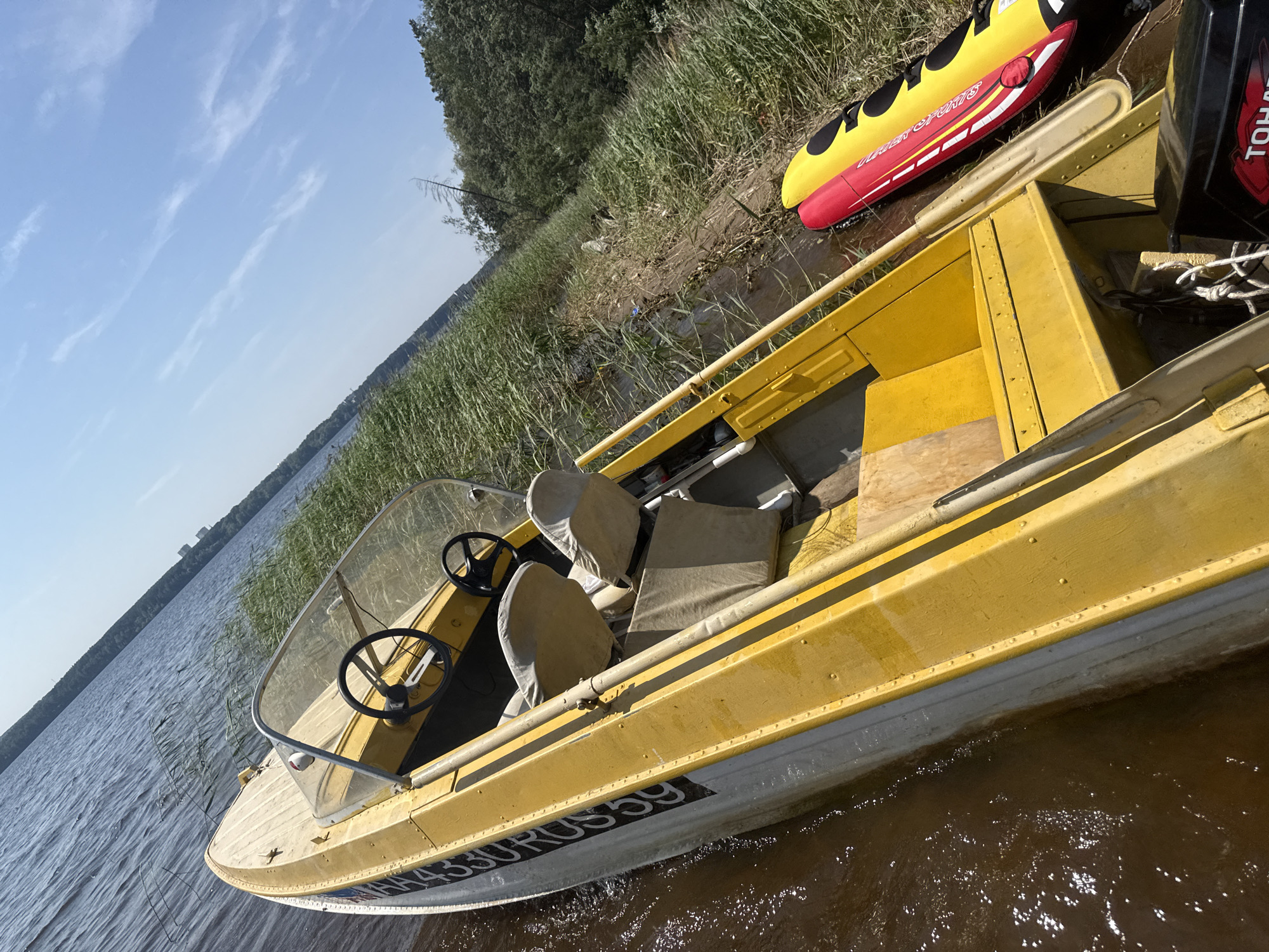 This is a medium-angle, outdoor shot of a yellow motorboat partially submerged in the shallows of a body of water, likely the Kama River or a reservoir near Chaikovsky, Russia. The boat is beached on a muddy shore lined with tall, green reeds. The boat's interior is visible, featuring two grey fabric seats, a wooden deck, and a steering wheel. A paddle rests inside the boat. To the right of the boat, on the shore, lies a red and yellow inflatable tube. The water is murky brown, and the sky above is clear blue with a few wispy white clouds. In the background, a dense line of green trees borders the water, and distant buildings are faintly visible on the horizon. The image is taken from a slightly elevated, tilted perspective, giving a dynamic view of the scene.