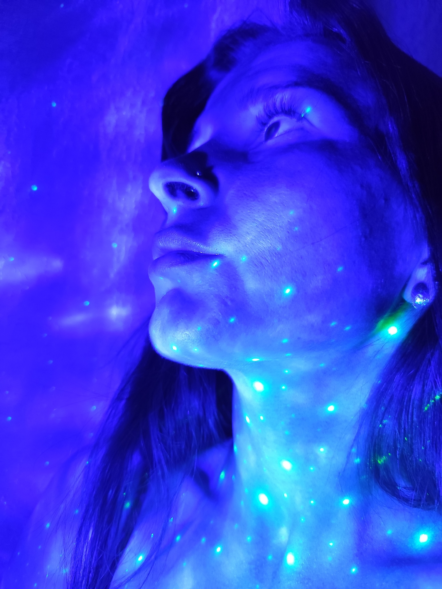 A close-up image depicts a person's face and neck illuminated with blue and green light. The person is facing left, with their gaze directed upwards. The light source appears to be projecting small points of light onto the skin. The individual has dark hair and fair skin, which takes on the blue hue of the light. There are no other discernible details about the surroundings to determine a location in Фоки, Russia.