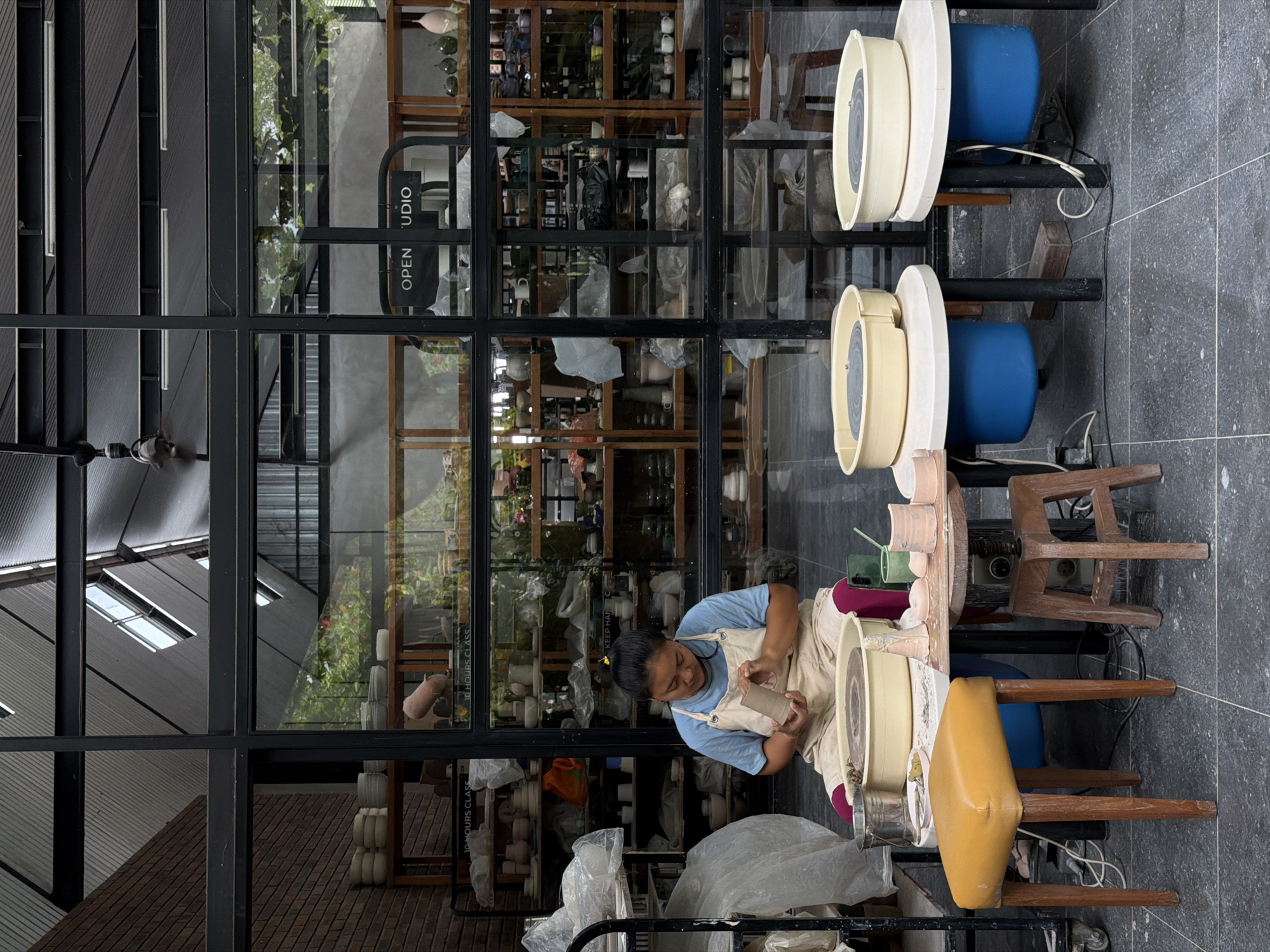 An outdoor pottery workspace in Singakerta, Indonesia, is depicted adjacent to a modern studio with a black-framed glass facade. A person, identified as female, with dark hair tied back, wears a light blue t-shirt and a beige apron, seated on a dark low stool. She is actively shaping a partially formed cylindrical clay object with both hands.
To her immediate left, a yellow pottery wheel with a lump of clay sits on the ground, accompanied by a metal bucket and a small white dish. To her right, a small wooden table holds several unfired clay pieces, including a small ornate white item, various rounded pinkish forms, and cylindrical segments, alongside green tools. The ground is paved with dark grey square tiles.
To the right of the central figure, a yellow upholstered stool with wooden legs is present. Further right, two additional pottery stations are set up, each consisting of a round white table with a blue base and a light-colored pottery wheel.
Through the glass facade of the studio, numerous wooden shelves are visible, densely packed with various ceramic items, predominantly in white, cream, and earthy tones. Signs such as "10 HOURS CLASS" and "OPEN STUDIO" are legible within the interior. The studio's architecture includes a brick wall section on the far left and an angled ceiling with exposed black beams and skylight-like openings.